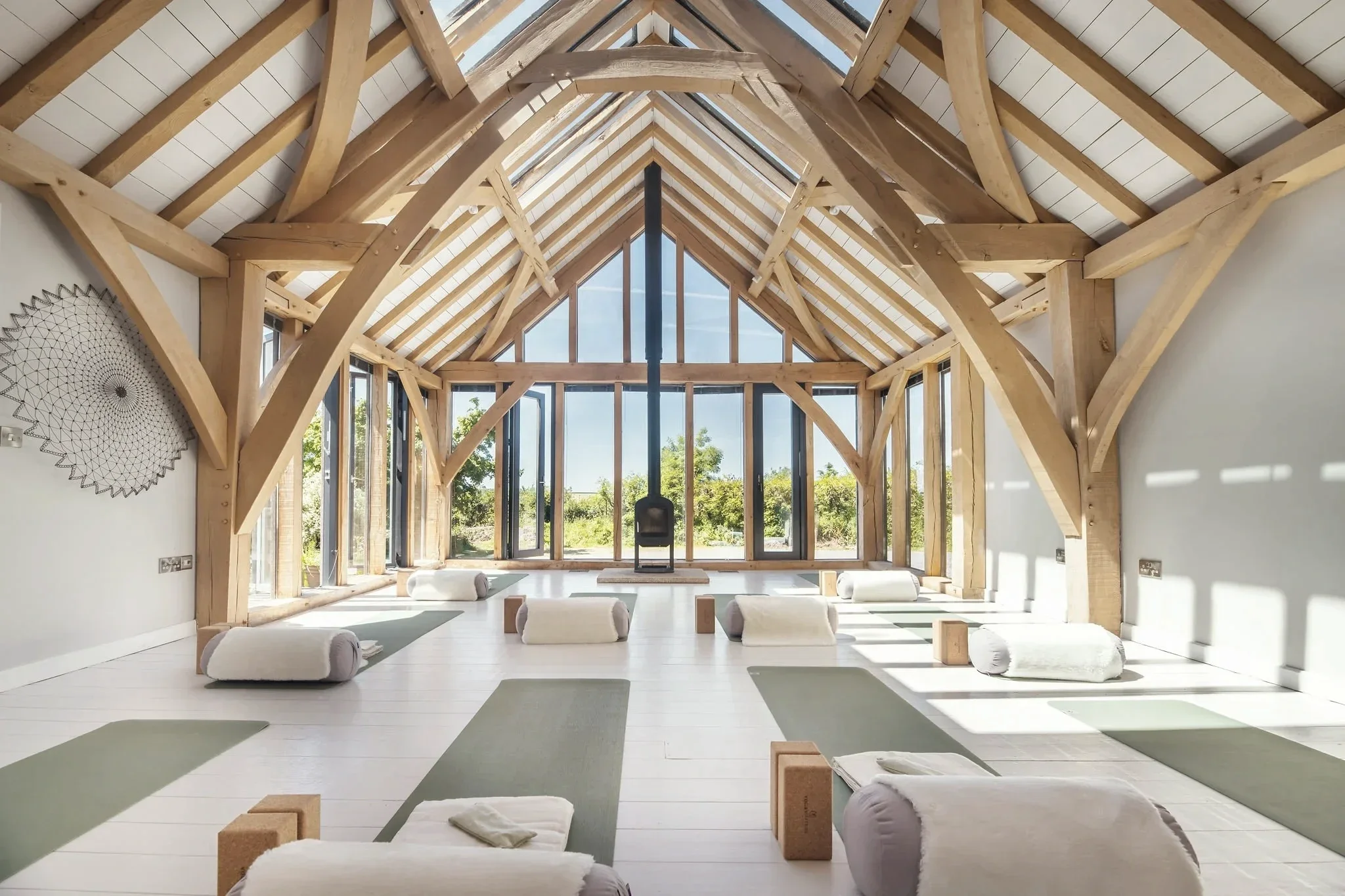A spacious yoga or meditation studio with wooden beams and large windows, furnished with yoga mats and bolster cushions, illuminated by natural light.