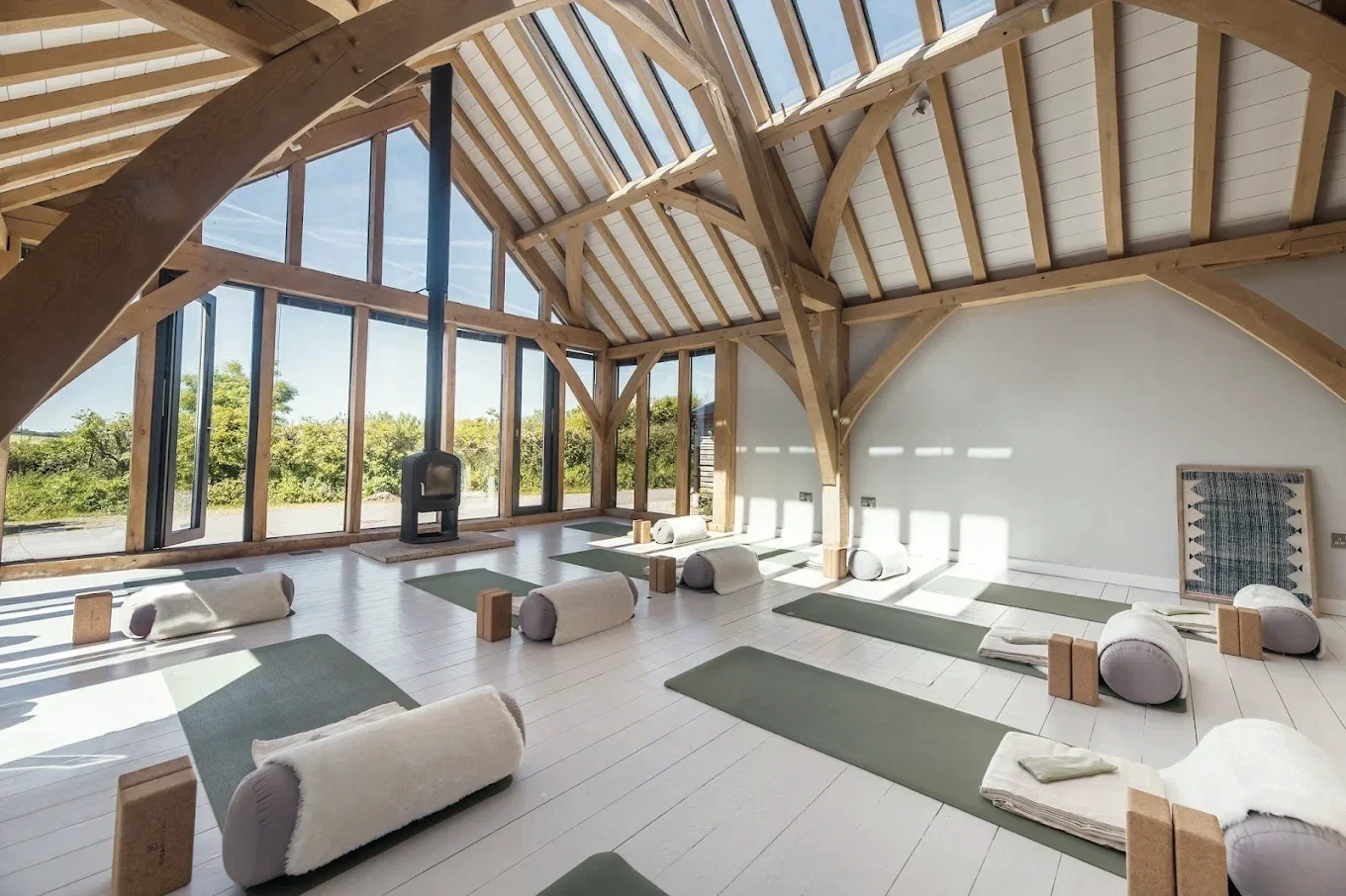 Interior of a yoga or meditation studio with wooden beams, large windows, and a fireplace. Mats and bolsters are arranged on the white wooden floor for practice.