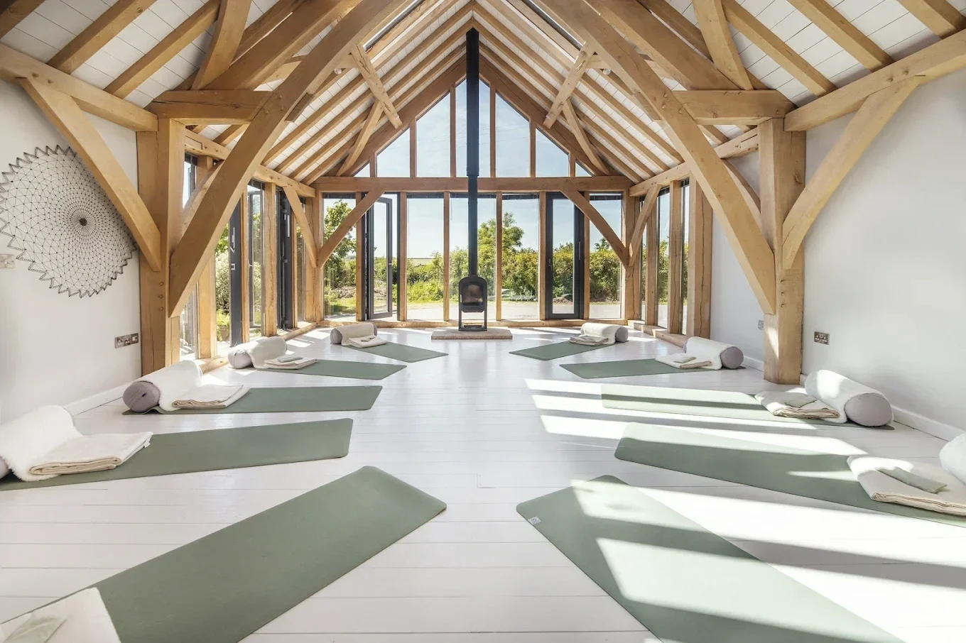 Sunlit yoga studio with wooden beams and large glass windows, green yoga mats and bolsters arranged on white floor for meditation or yoga practice.