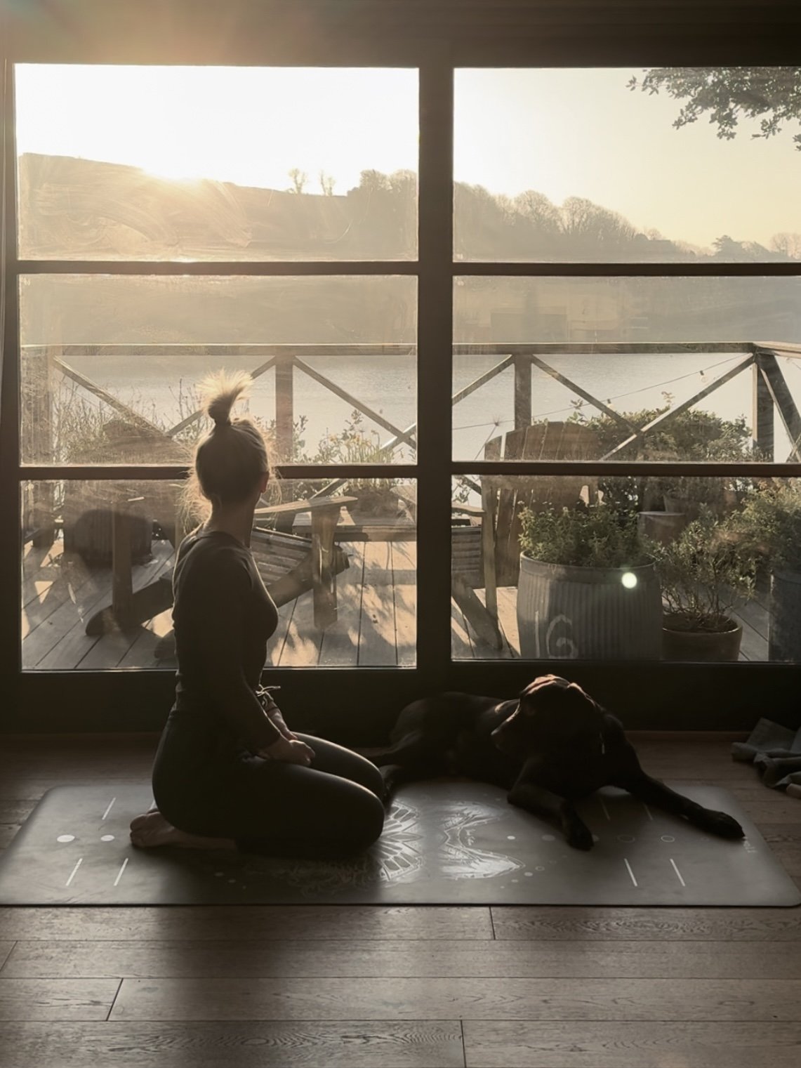 A woman practicing yoga on a mat inside, sitting in a kneeling pose, with a large dog lying nearby, looking out a large window at a lakeside sunset with outdoor furnishings and plants outside.
