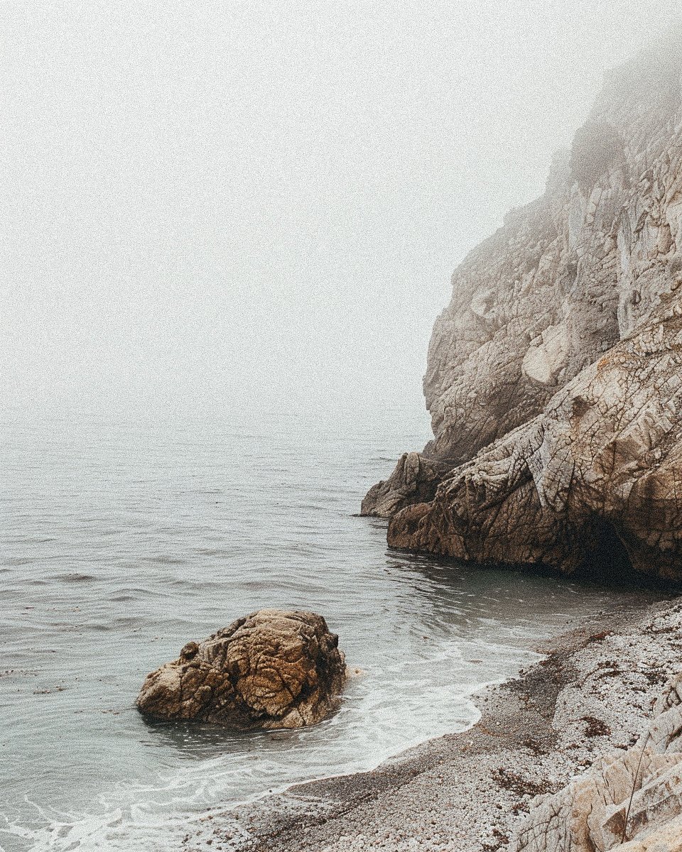 Foggy rocky coastline with a visible cliff, a large rock in the water, and gentle waves reaching a small pebble beach.
