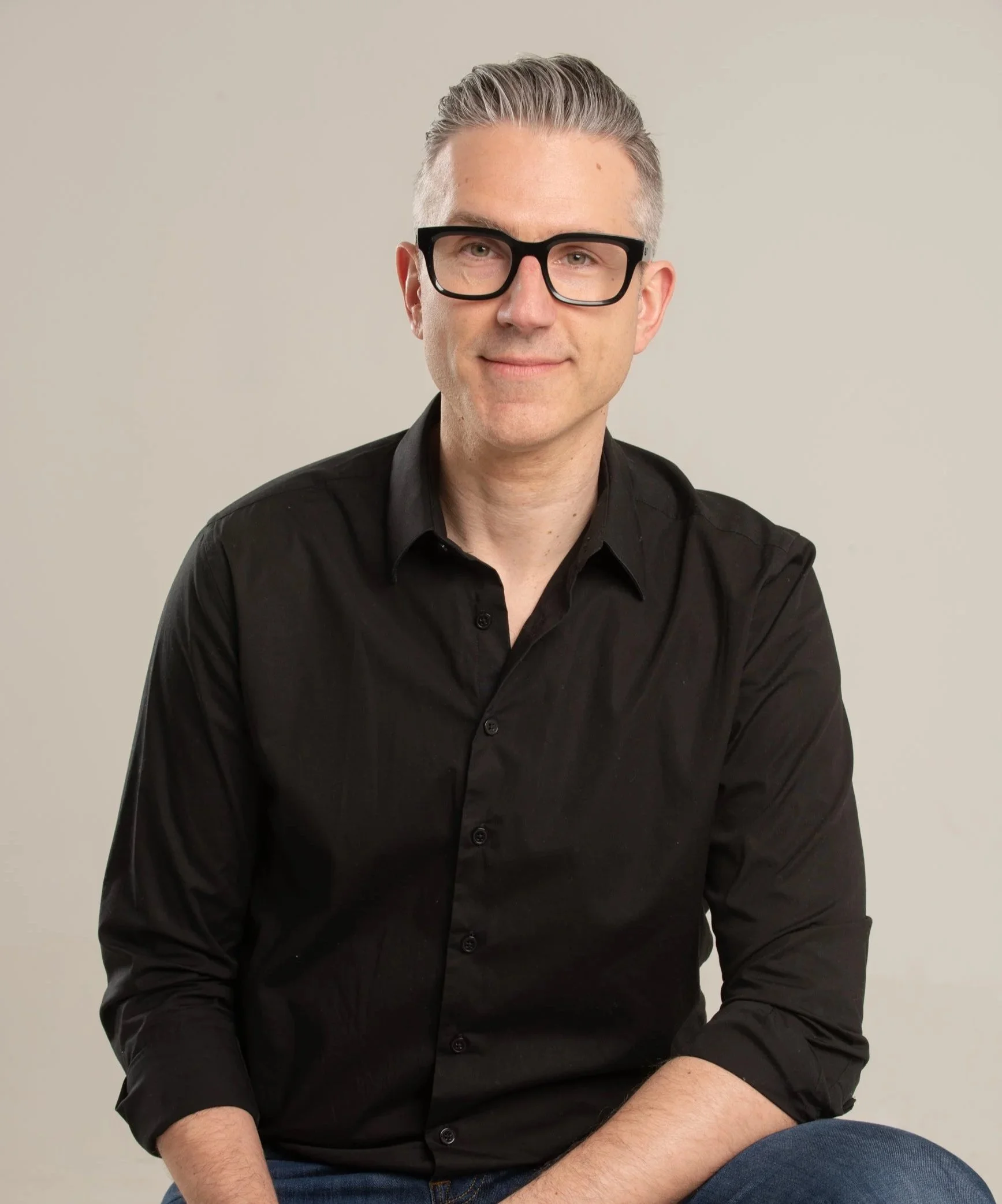 Stéphane Protat, cofondateur de RéseauPsy et psychologue, portant une chemise noire et des lunettes épaisses, avec des cheveux gris, souriant légèrement, pose devant un fond neutre.