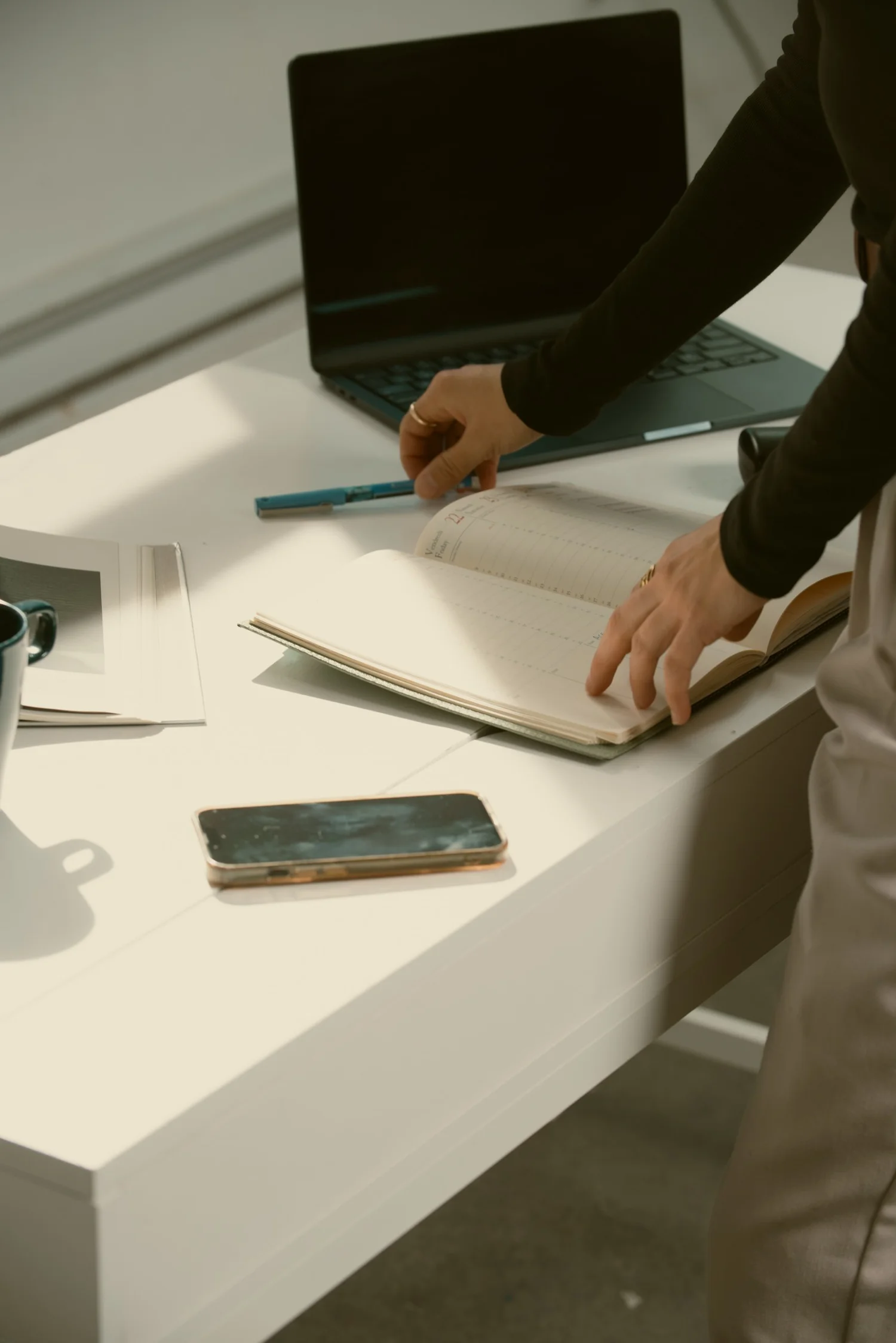 Personne manipulant un agenda ouvert sur un bureau blanc avec un ordinateur portable, un smartphone et une tasse