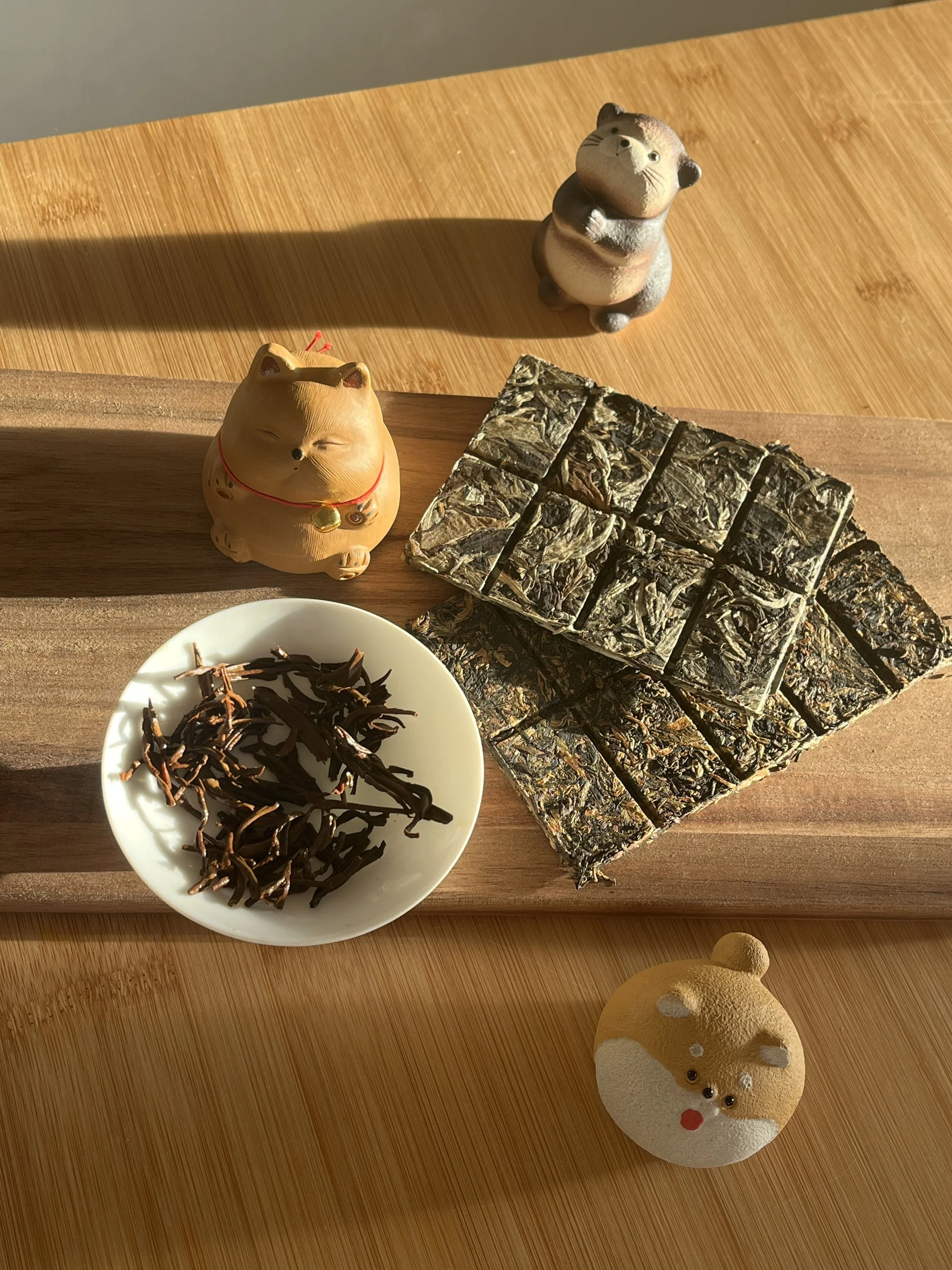 Auf einem Holztisch sind getrocknete Teeblätter, eine kleine Keramikfigur eines glücklichen Katzen-Glücksbringers, ein Stück zusammengepresster Teeblock, zwei kleine Figuren eines Otters und eines Hundes.