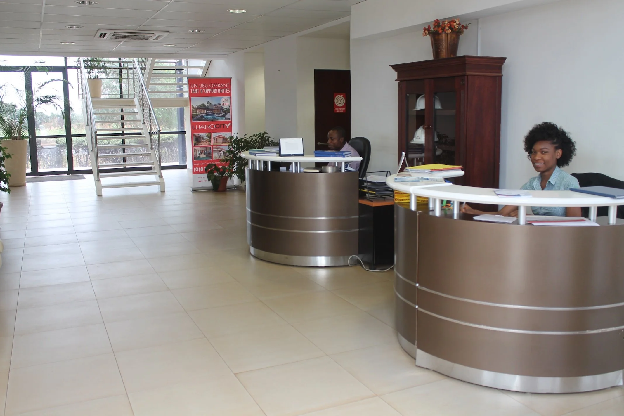 Office reception area with two women working at curved desks, potted plants, staircase, promotional banner, and a large window at the commercial offices, Luano City.