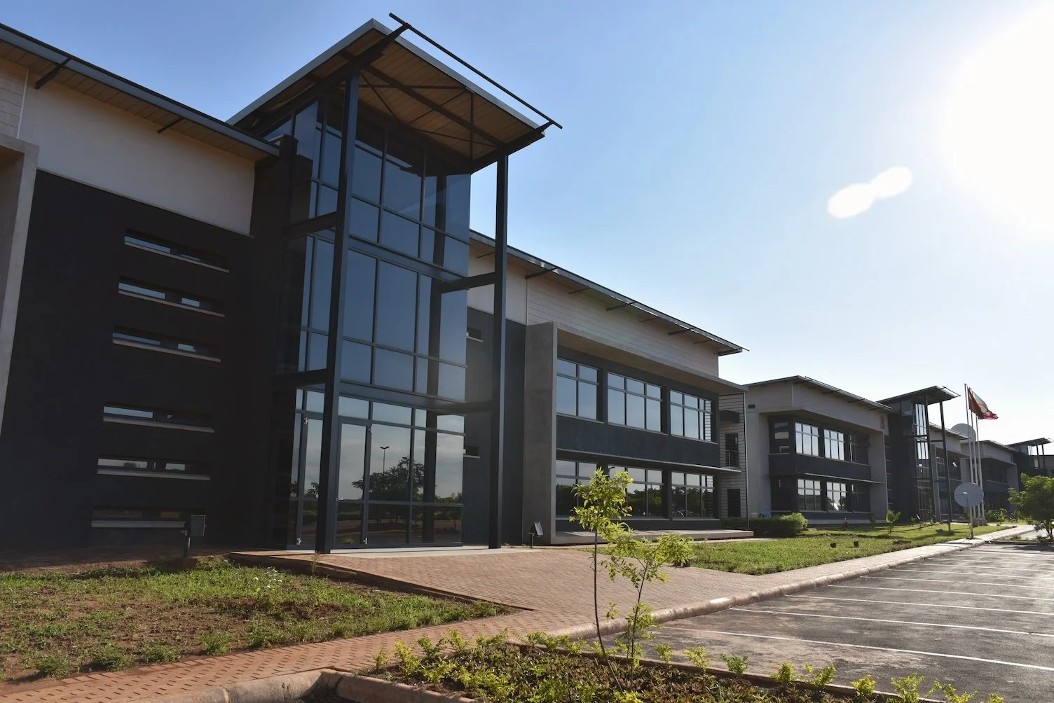 Modern office building with glass walls and parking lot, small trees, American flag, sunny day at the commercial offices, Luano City.