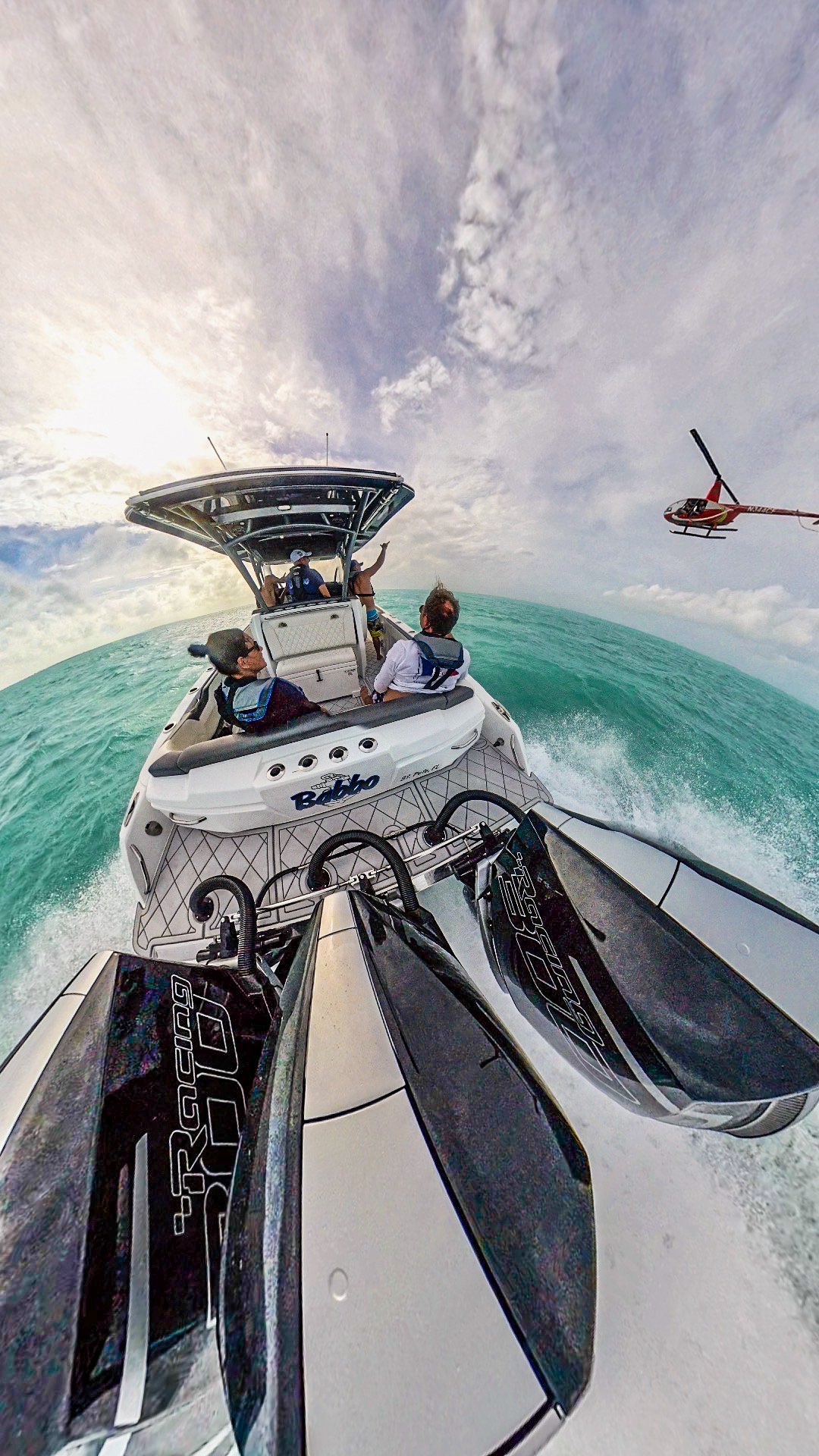 A boat with people on it is moving through the water, with a helicopter flying nearby in the sky.