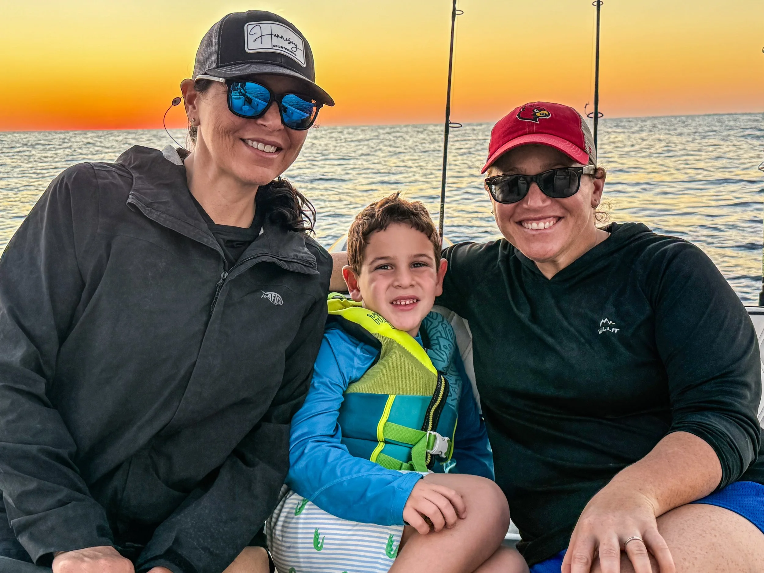 Three people, an adult woman, a young boy, and another adult woman, smiling and posing for a photo on a boat during sunset with a calm ocean in the background. They are dressed in casual outdoor clothing and wearing sunglasses and hats.  After fishin