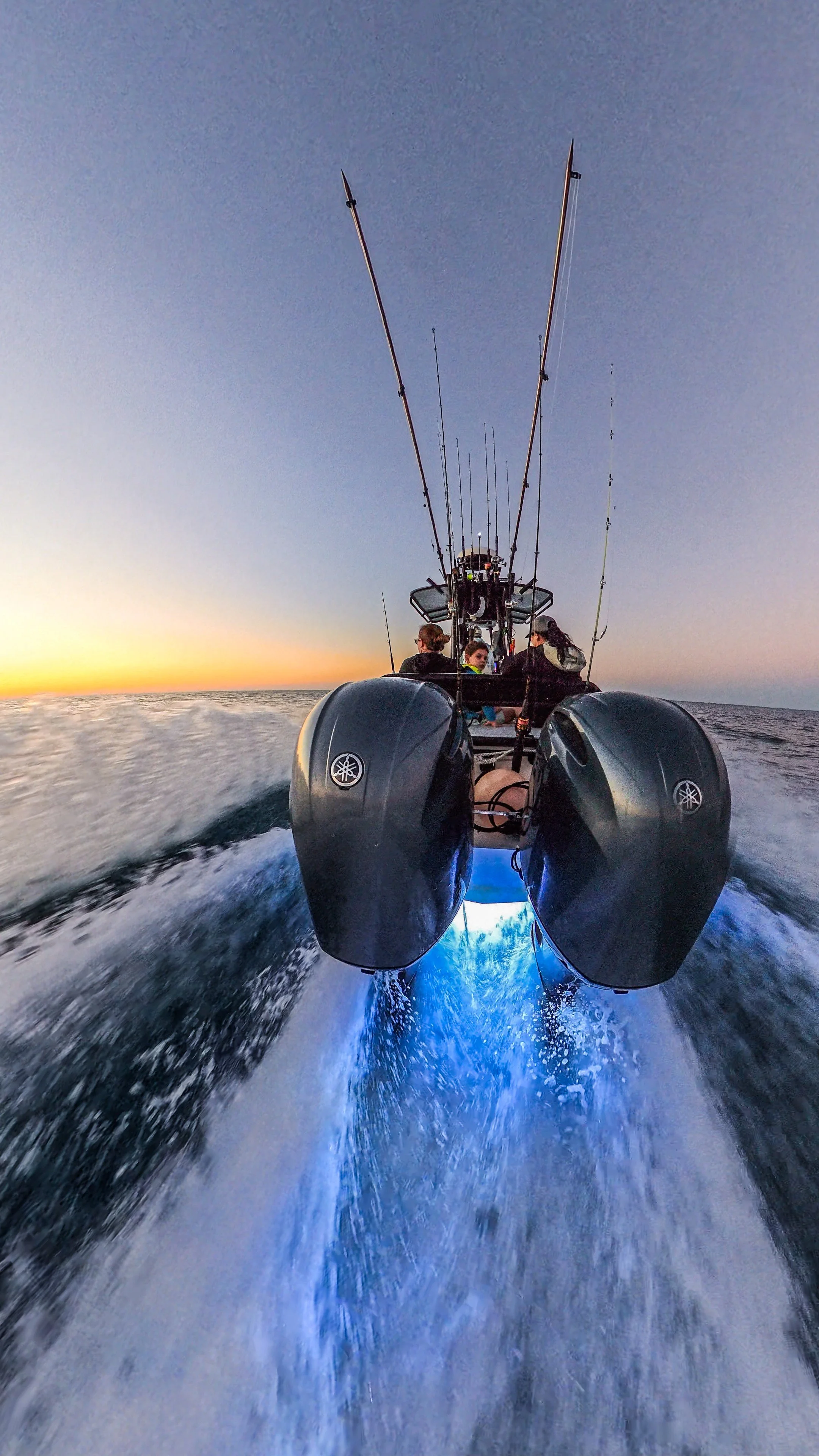 A boat speeding through the water at sunset with people on board and fishing rods attached.