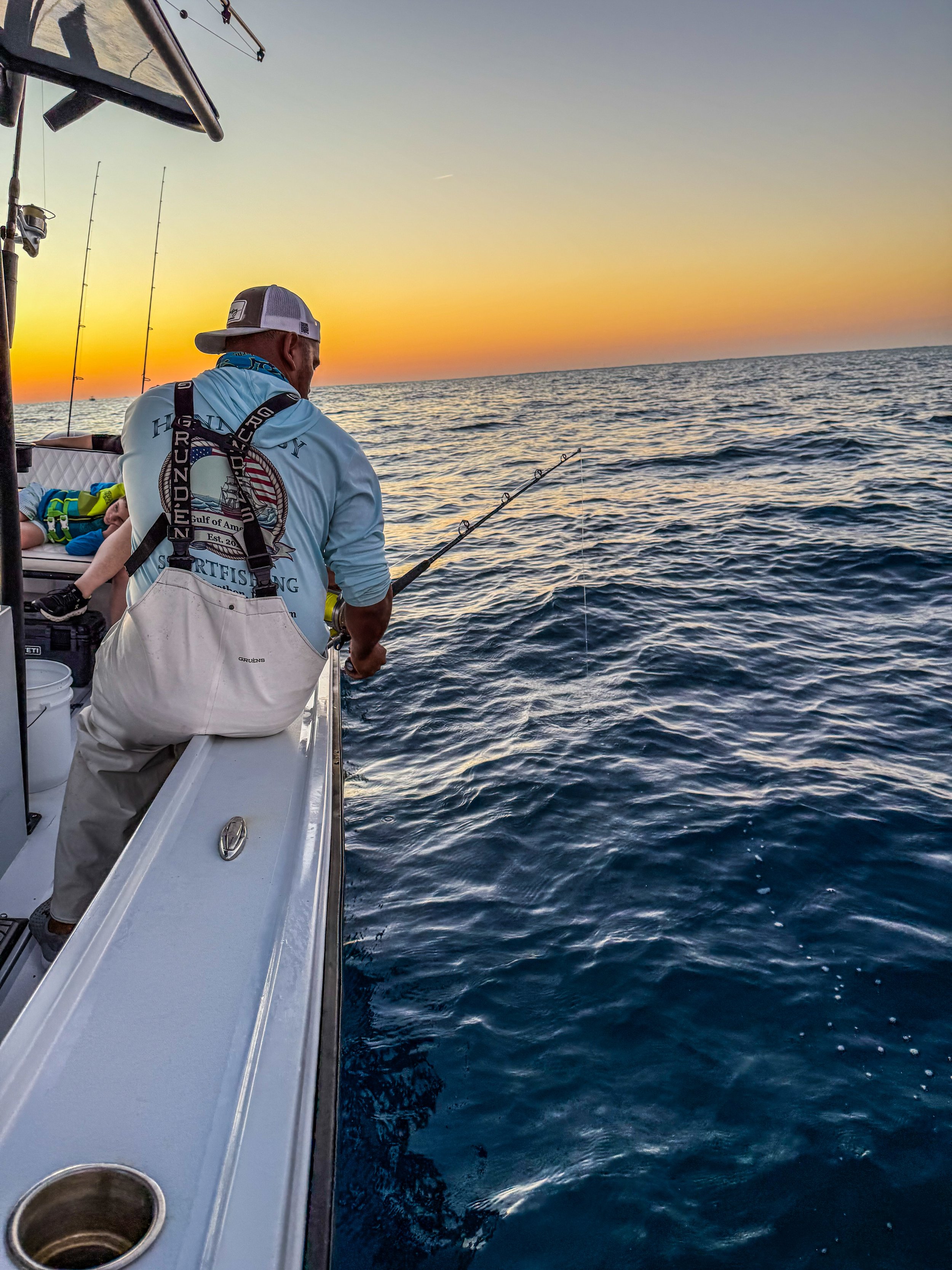 A man with a white cap and fishing gear on a boat at sunset, fishing in the ocean.