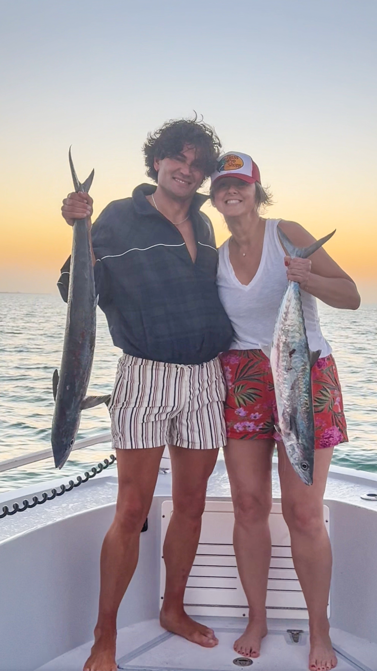 a man and women stand on a boat holding giant fish that they just caught at sunset over the ocean.