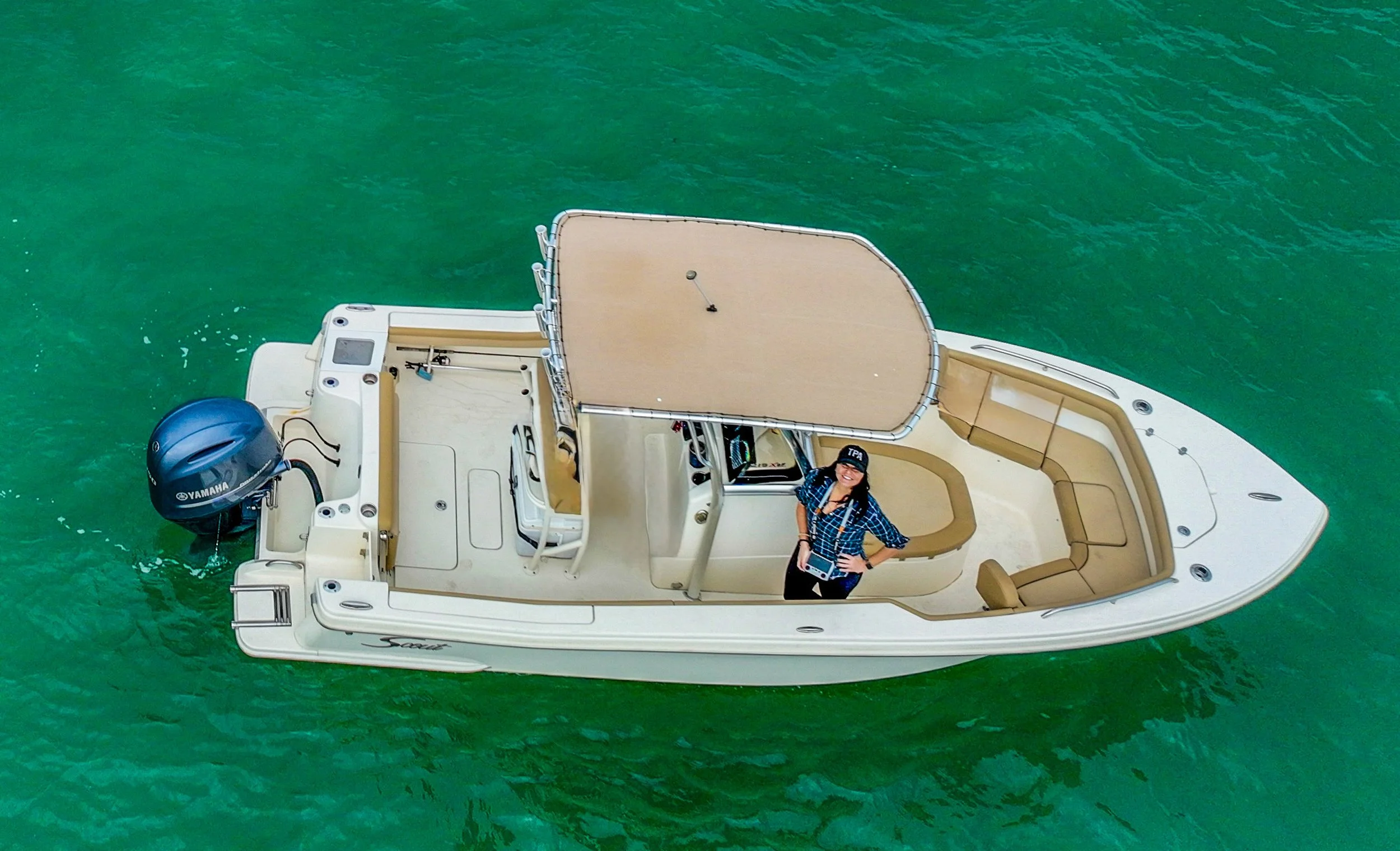 A woman standing on a beige and white boat with a captain's chair, steering wheel, and an outboard motor, floating on green water, holding a tablet and smiling.