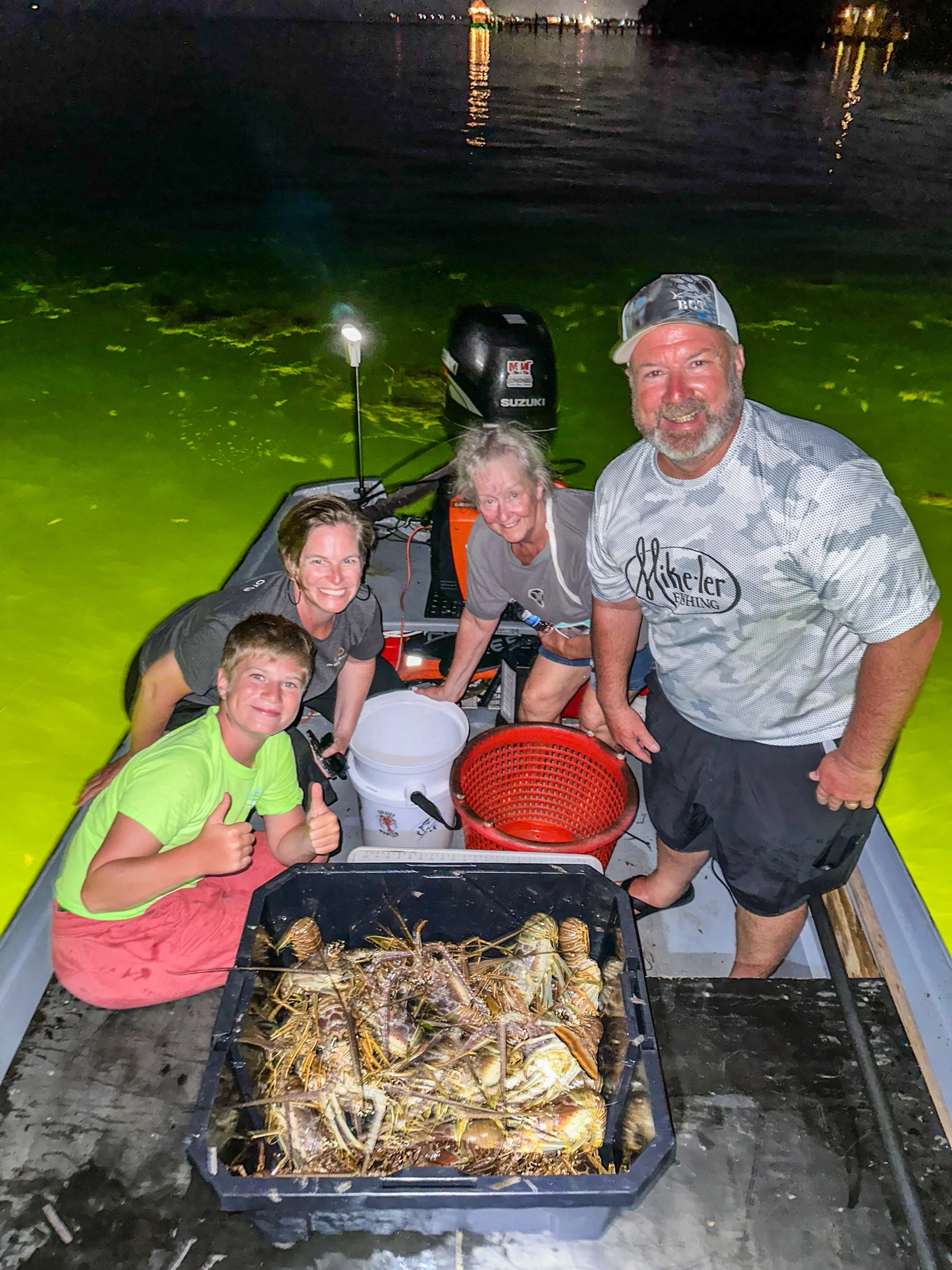 A group of five people on a boat at night, with a large catch of lobsters in a tray at the front. The background shows a body of water with reflections of city lights.