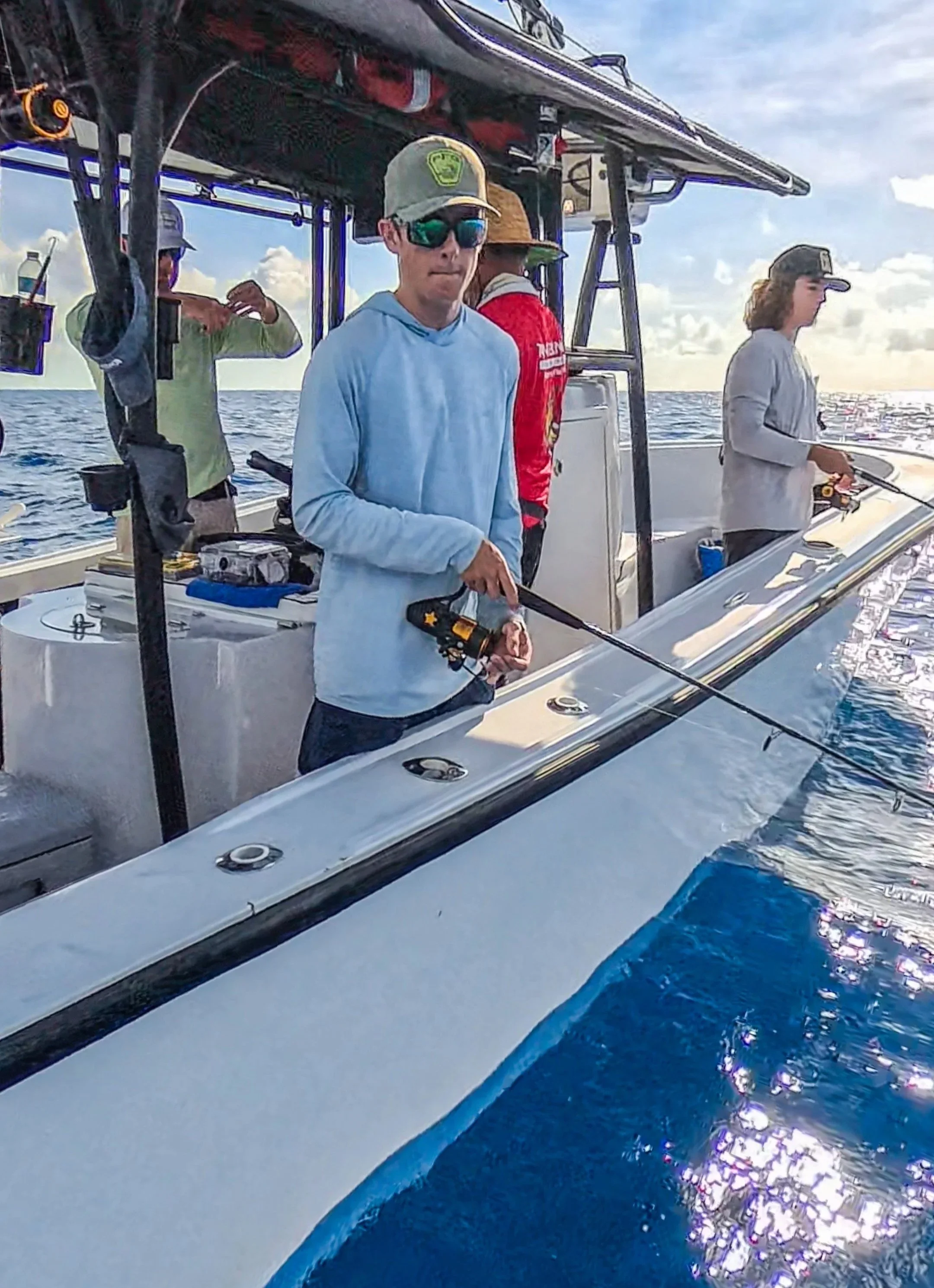 People fishing on a boat in the ocean, with a young man in sunglasses and a baseball cap holding a fishing rod, and others in the background preparing fishing gear under a partly cloudy sky.