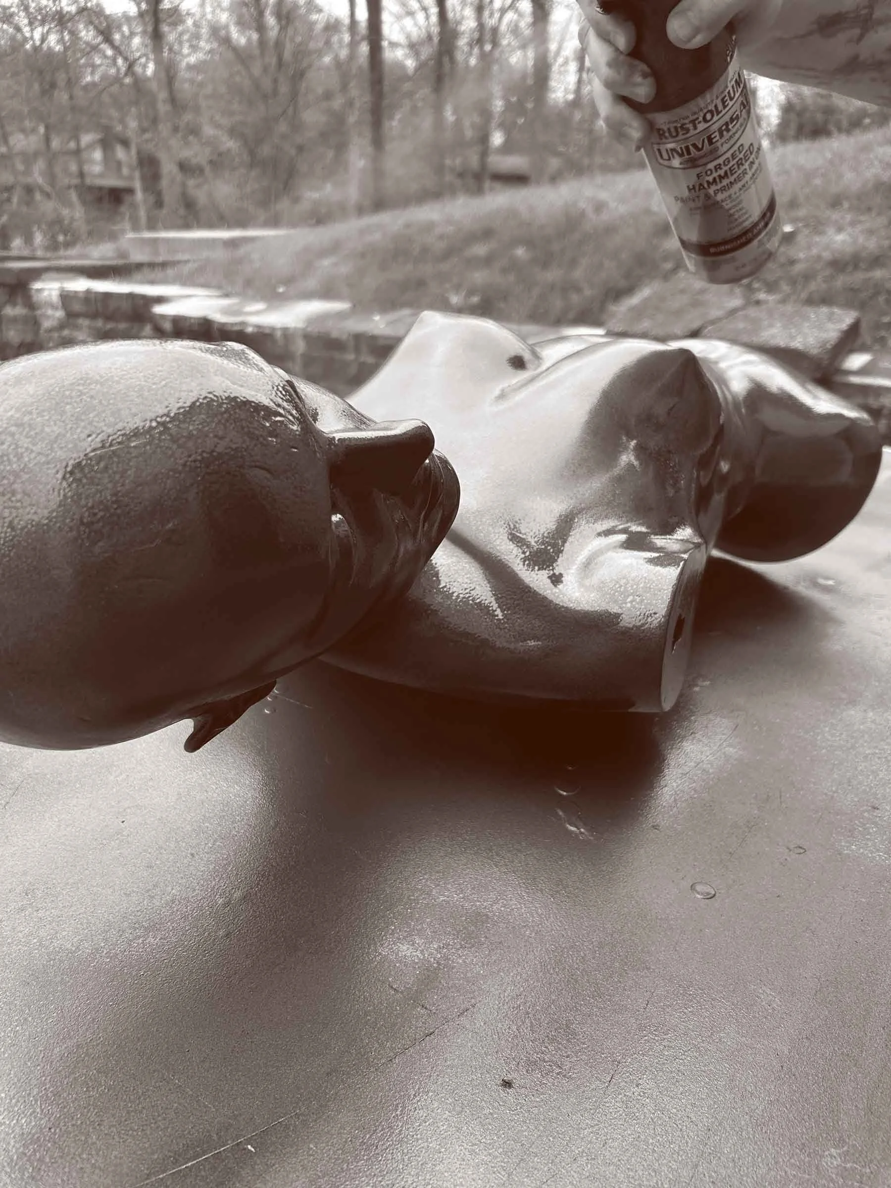 A person wearing gloves holds a small container of hammered spray paint above a ceramic torso sculpture, which is outdoors on a concrete surface with trees in the background.