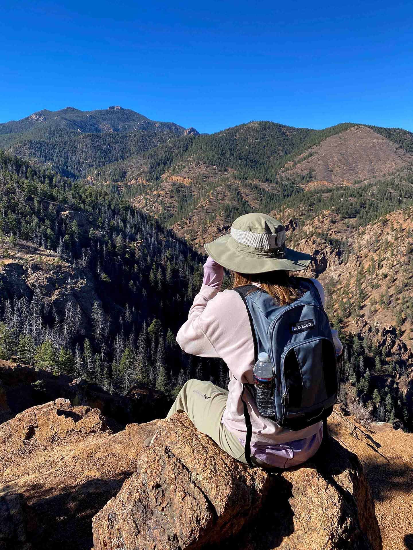 Person sitting on rocky terrain overlooking a mountain range with dense pine forests and clear blue sky, wearing a wide-brimmed hat, a backpack, and holding a water bottle.
