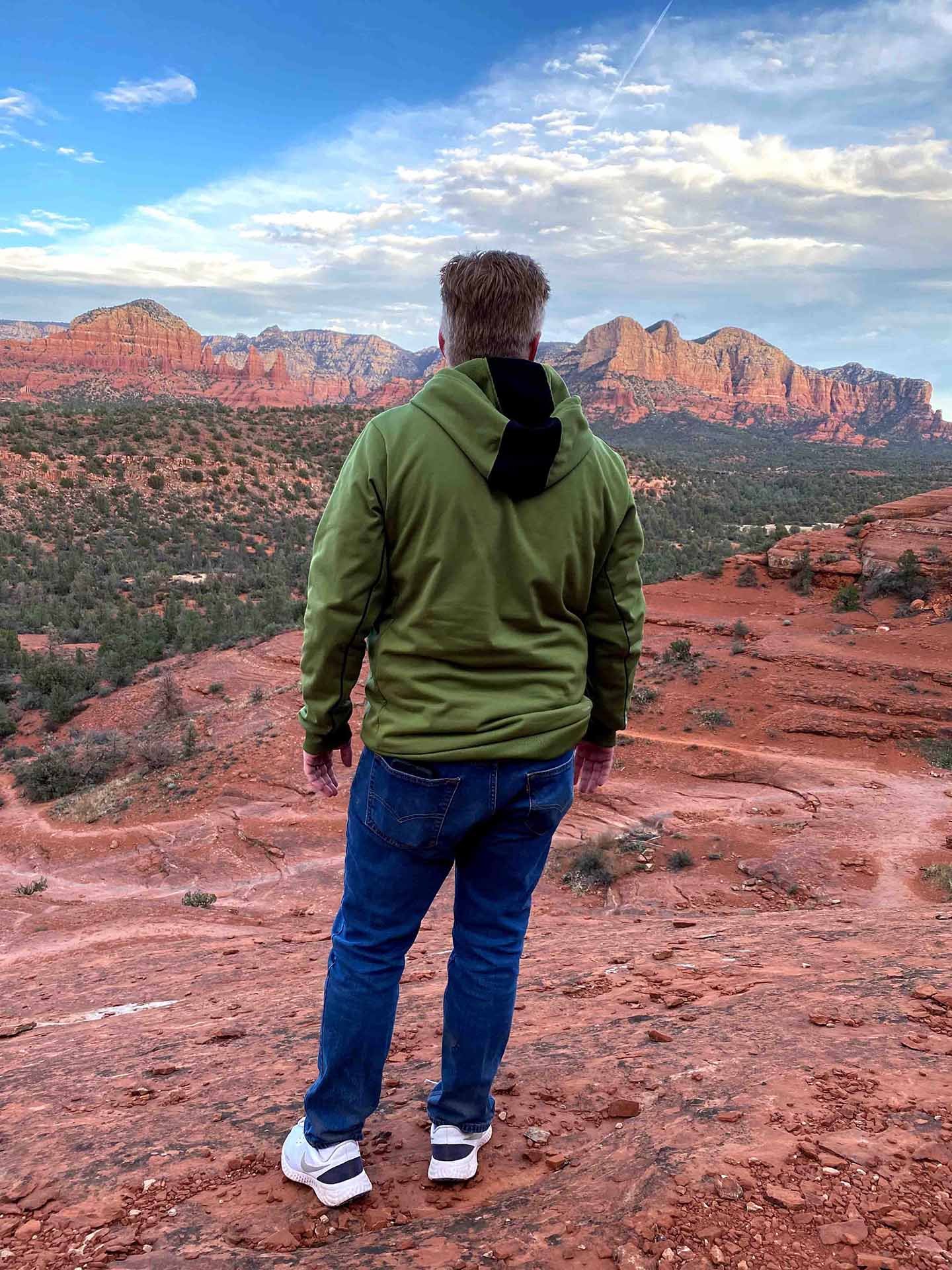 A person with blonde hair wearing a green jacket and blue jeans stands on a reddish rocky landscape, looking at red rock formations and mountains in the distance, under a partly cloudy sky.
