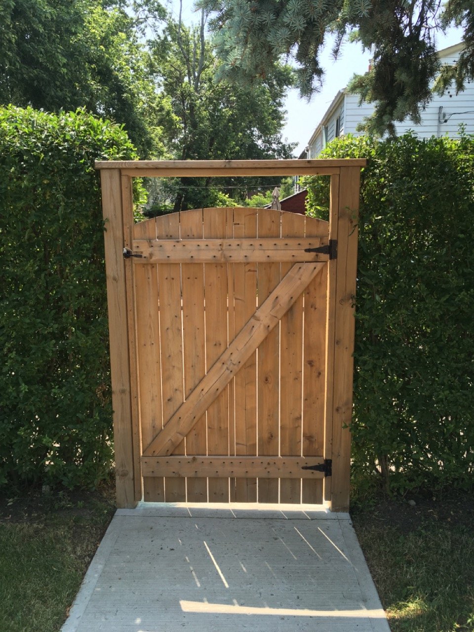Wooden garden gate with metal hinges, surrounded by green hedges and trees, on a concrete path.