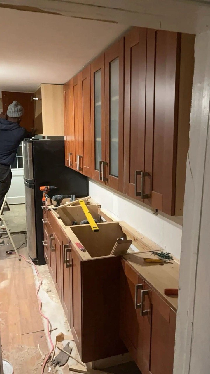 Kitchen under renovation with wooden cabinets, an open drawer, construction tools, and a person standing on a ladder.