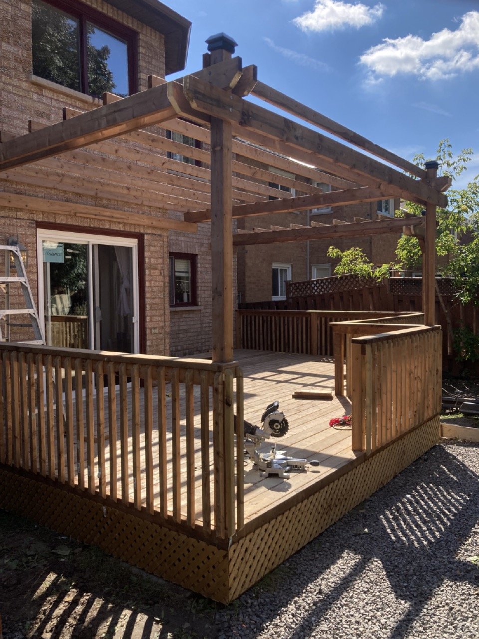 Wooden deck with pergola under construction, attached to a brick house with sliding glass door and a circular saw on the deck.