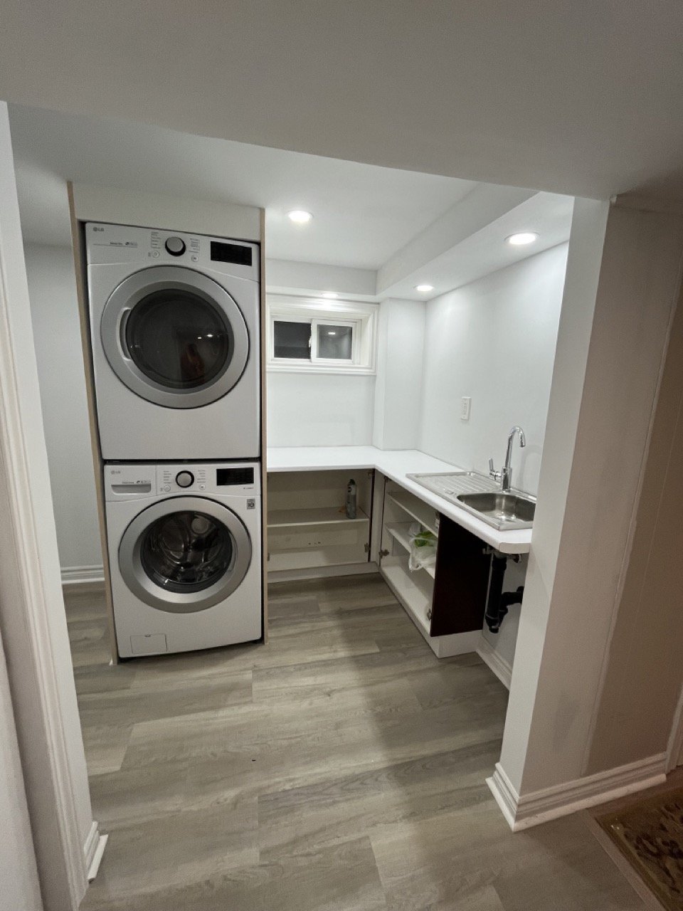 Modern laundry room with stacked washer and dryer, white cabinetry, sink, and small window.