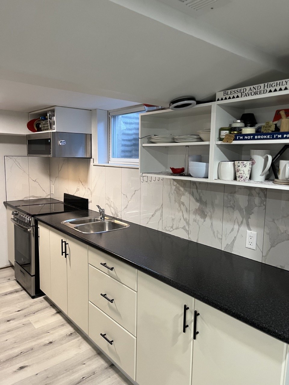 Modern kitchen with black countertop, stainless steel stove, white cabinets, open shelving with dishes and mugs, marble backsplash, and a small window.