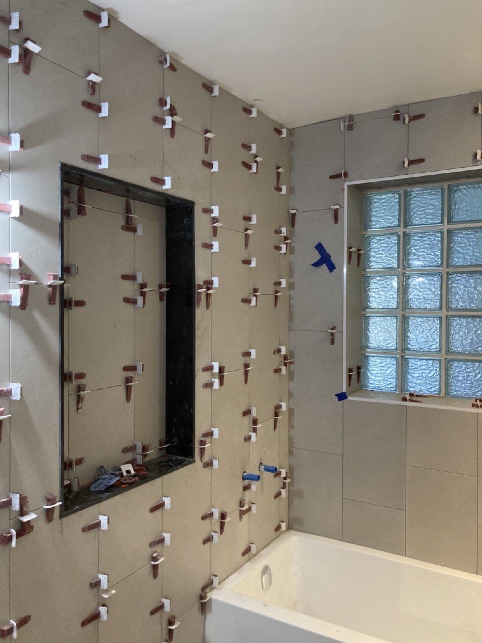 Bathroom under renovation with tile spacers on walls, beige tiles, a bathtub, and glass block window.