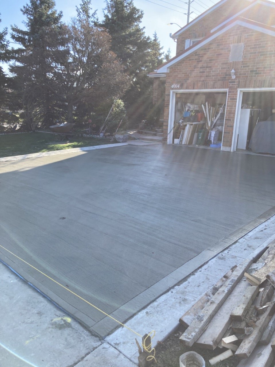 Freshly poured concrete driveway in front of a brick house with an open garage displaying various items, surrounded by trees and sunlight.