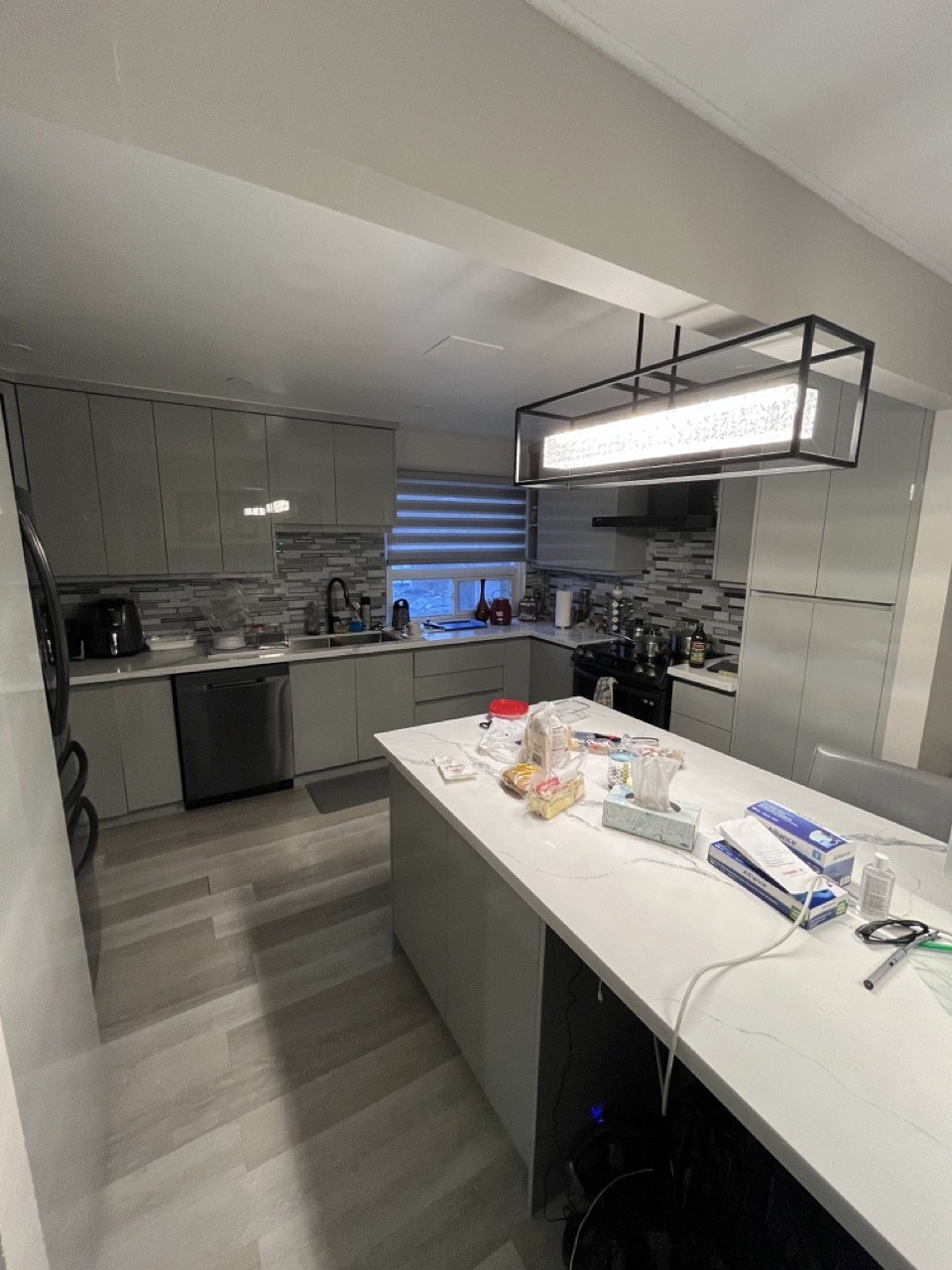 Modern kitchen with gray cabinets, a large island, and a contemporary light fixture. The countertop has various items including food packages and cleaning supplies. The backsplash features a tiled design and there are stainless steel appliances like