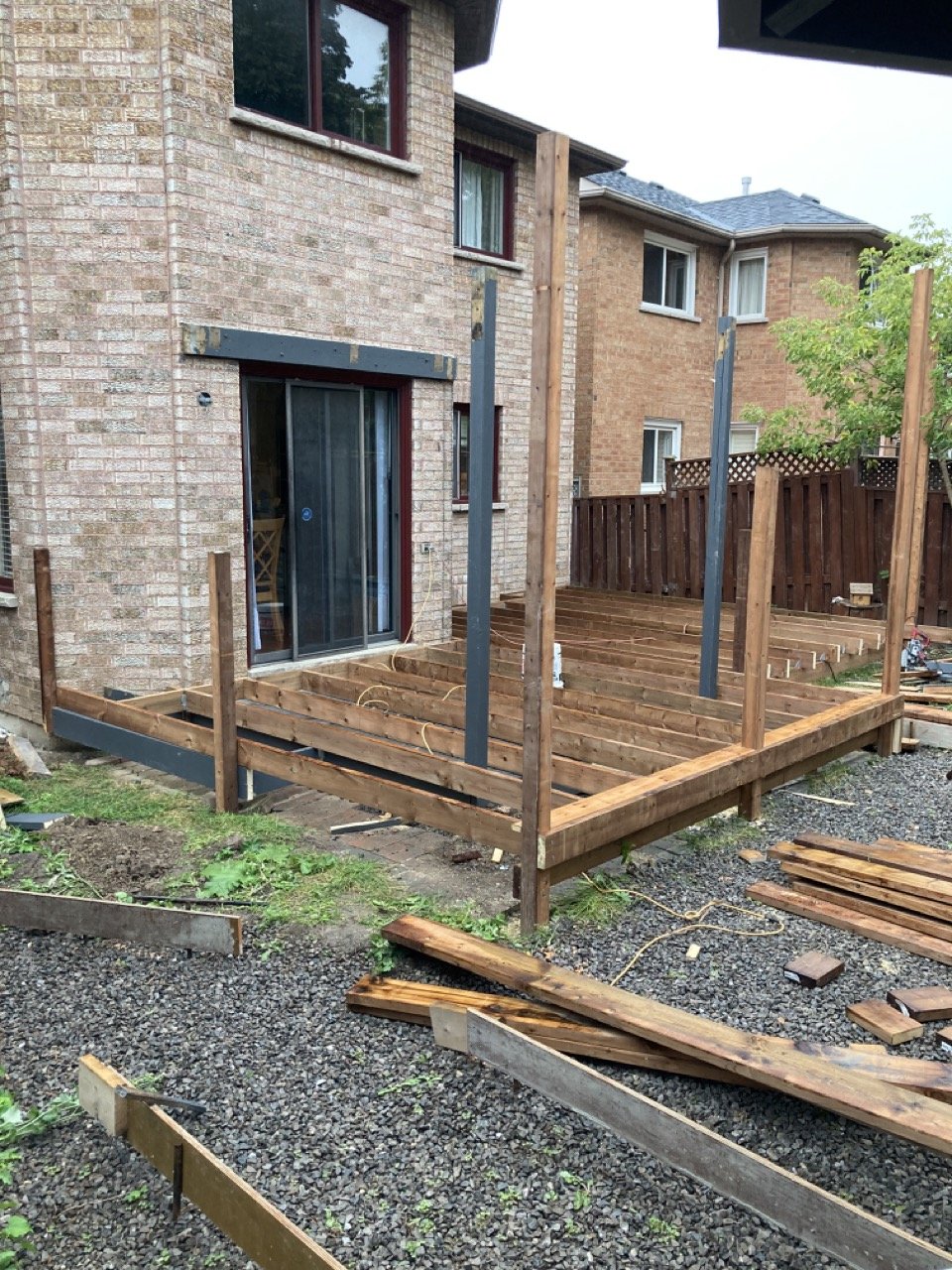Partially constructed wooden deck attached to a brick house with missing planks and posts. Nearby are scattered lumber pieces and gravel.