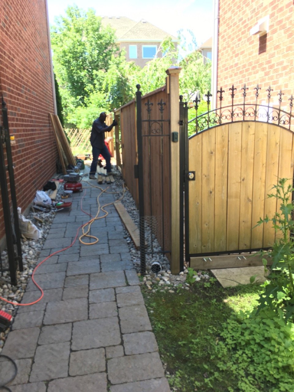 Person installing a wooden fence between two brick buildings on a sunny day, with gardening tools and supplies nearby.