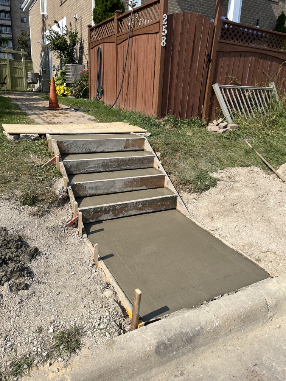 Freshly poured concrete steps and walkway under construction next to a brick house and wooden fence.