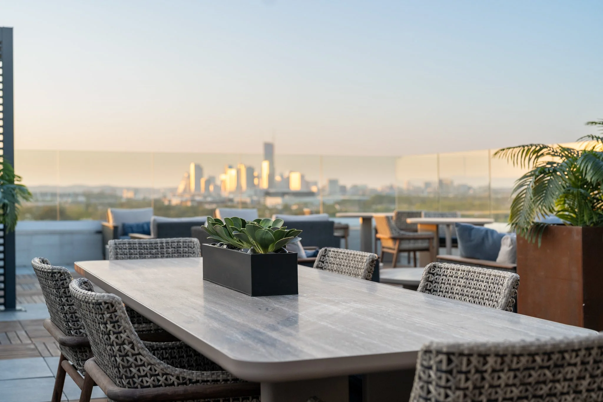 	
Architectural Photography featuring rooftop Patio of Zoey Apartments in Austin Texas. Outdoor rooftop dining area with a large table, wicker chairs, potted plants, and a city skyline view in the background during daytime.