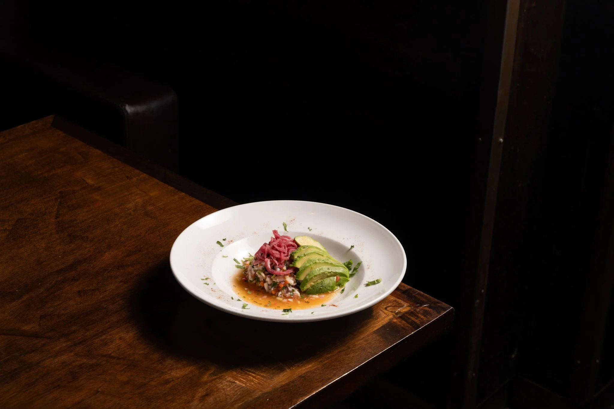 A white plate with thinly sliced avocado, a small portion of pink shredded meat or seafood, on top of a bed of chopped vegetables or grains, garnished with herbs and spices, with a light sauce or broth underneath, placed on a wooden table.