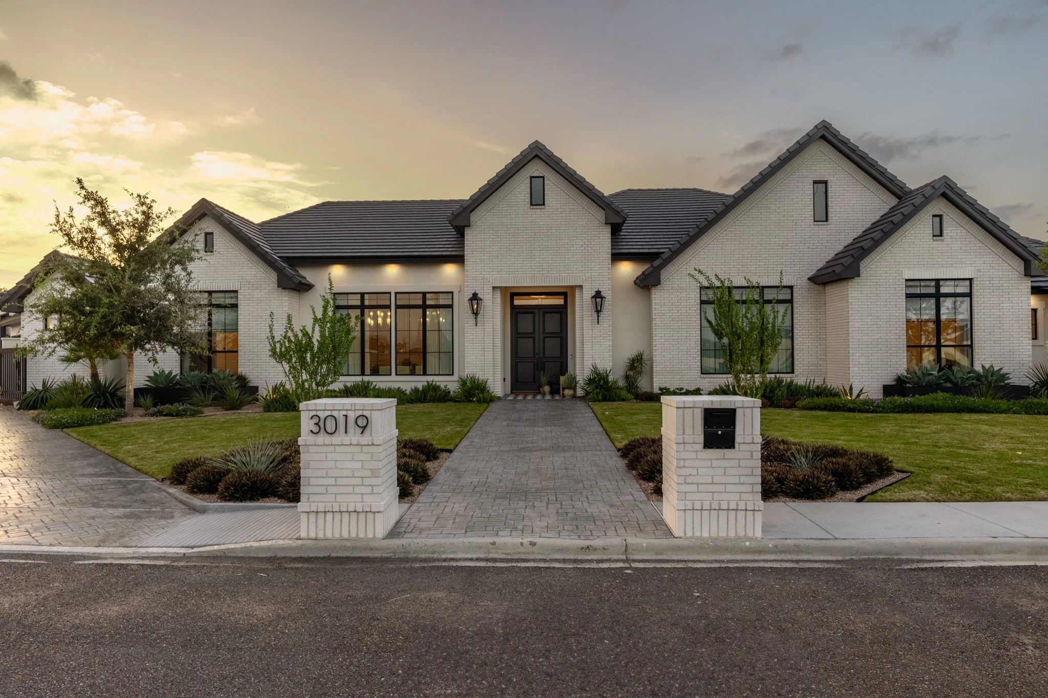 Front view of a modern white brick house with dark shutters and a central black door, a paved walkway leading to the entrance, green lawn, landscaped plants, and sunset sky.