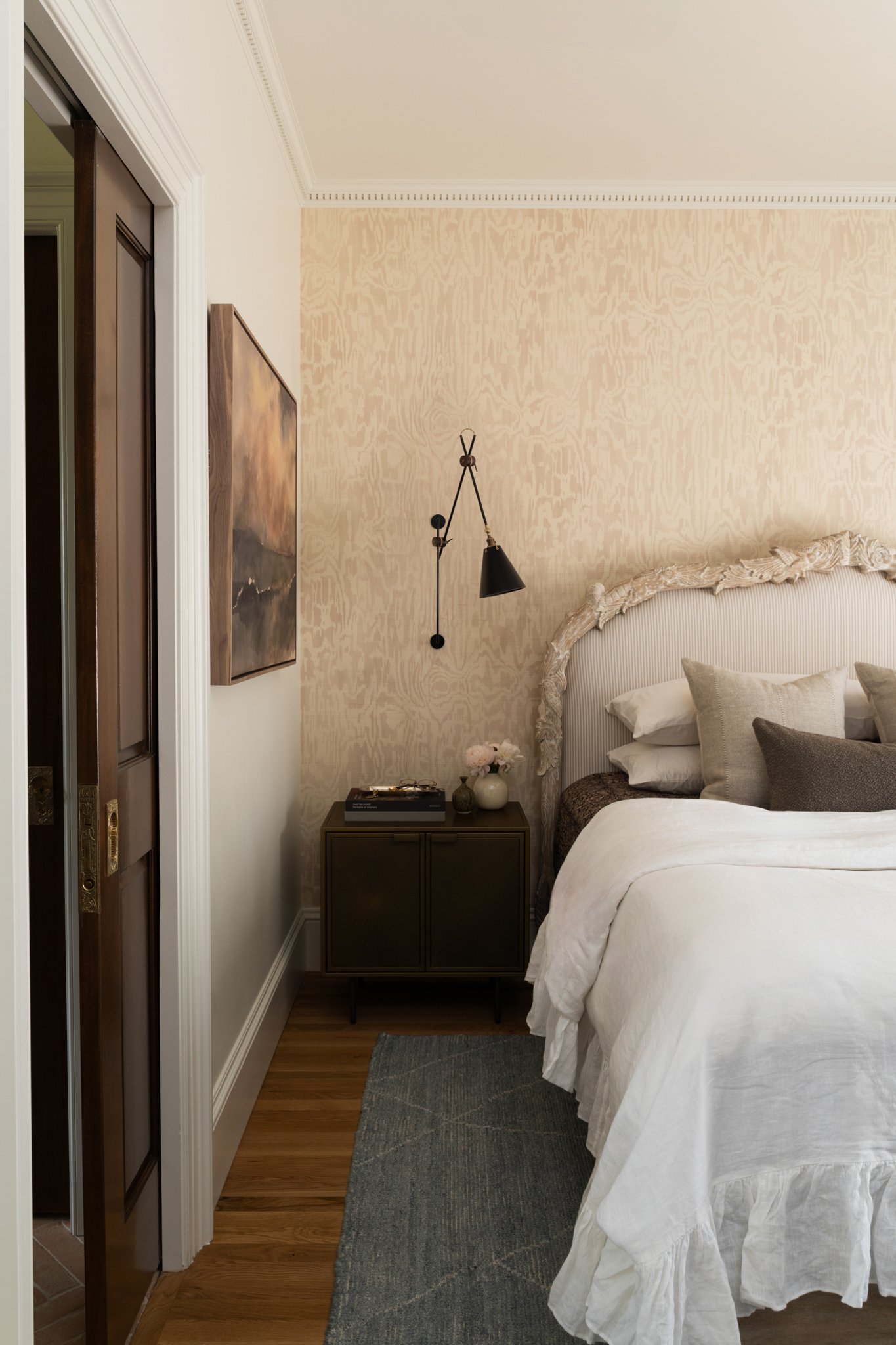 A cozy bedroom corner with a bed featuring cream-colored pillows and ornate headboard, a nightstand with decorative items, a wall-mounted black lamp, and soft beige textured wallpaper.