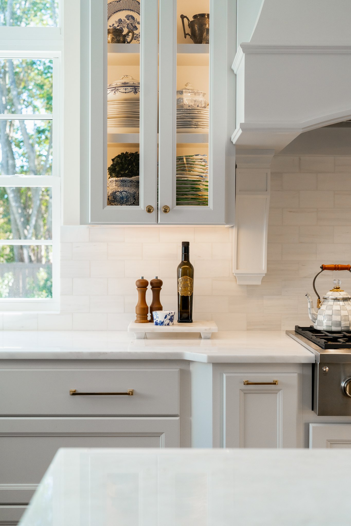 White kitchen with open cabinetry displaying dishes, some with blue patterns, and small potted plant. Counter with a bottle of olive oil, two wooden salt and pepper shakers, and a small dish. Stovetop with a silver kettle, and a window showing trees 