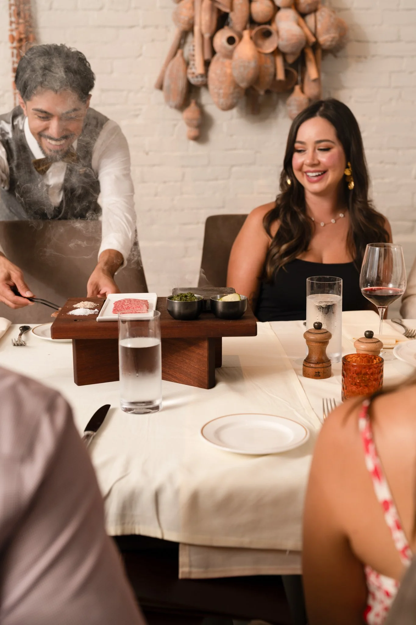 Restaurant Photography of Dean's Steakhouse at Kimpton Santo in San Antonio enjoying a hot plate meal at a restaurant with a chef preparing food, a woman in a black dress smiling, glasses of water and wine, and a rustic decor in the background.