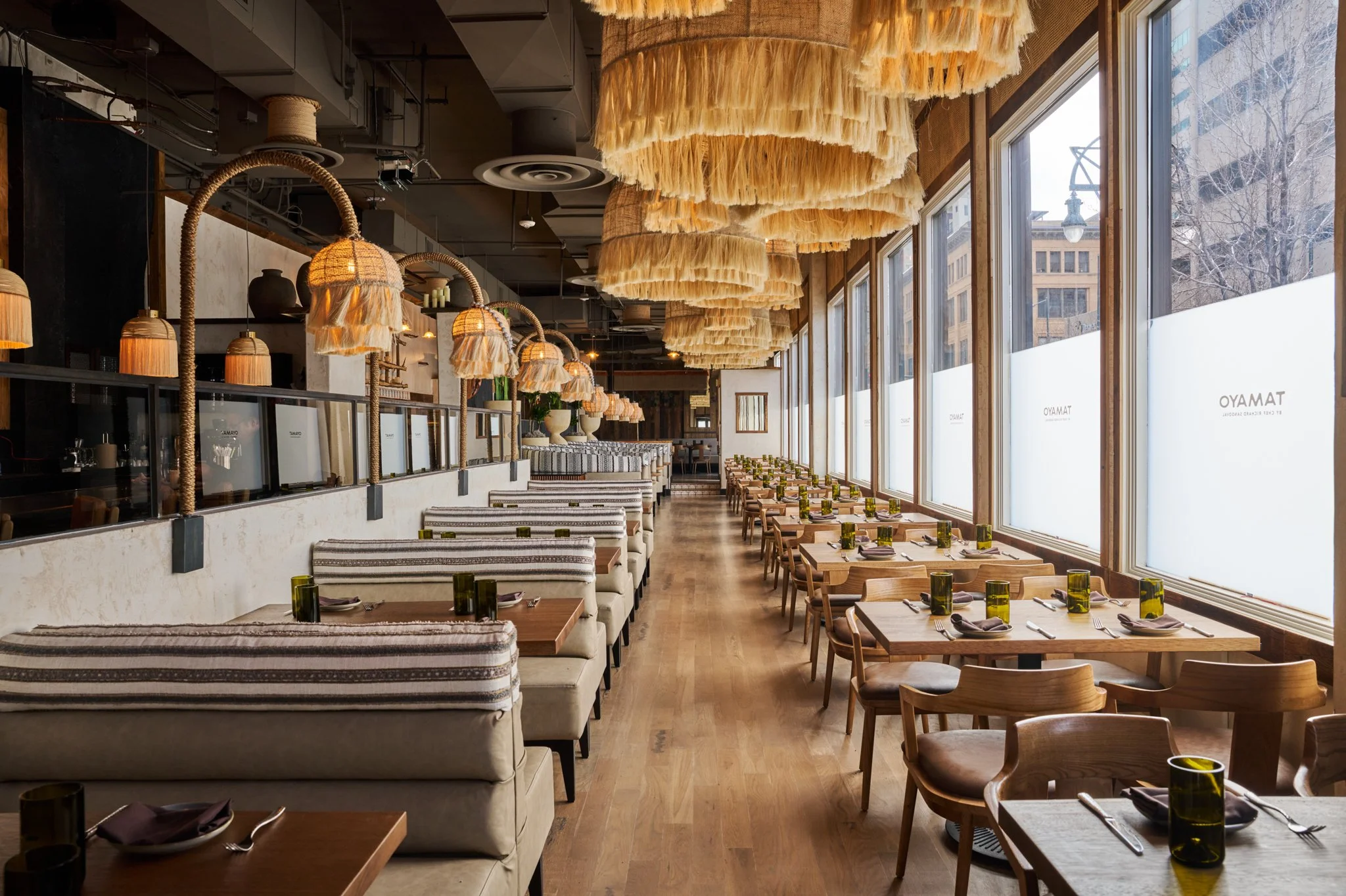 A modern restaurant interior with wooden tables and chairs, large windows with frosted glass, and hanging woven pendant lights.