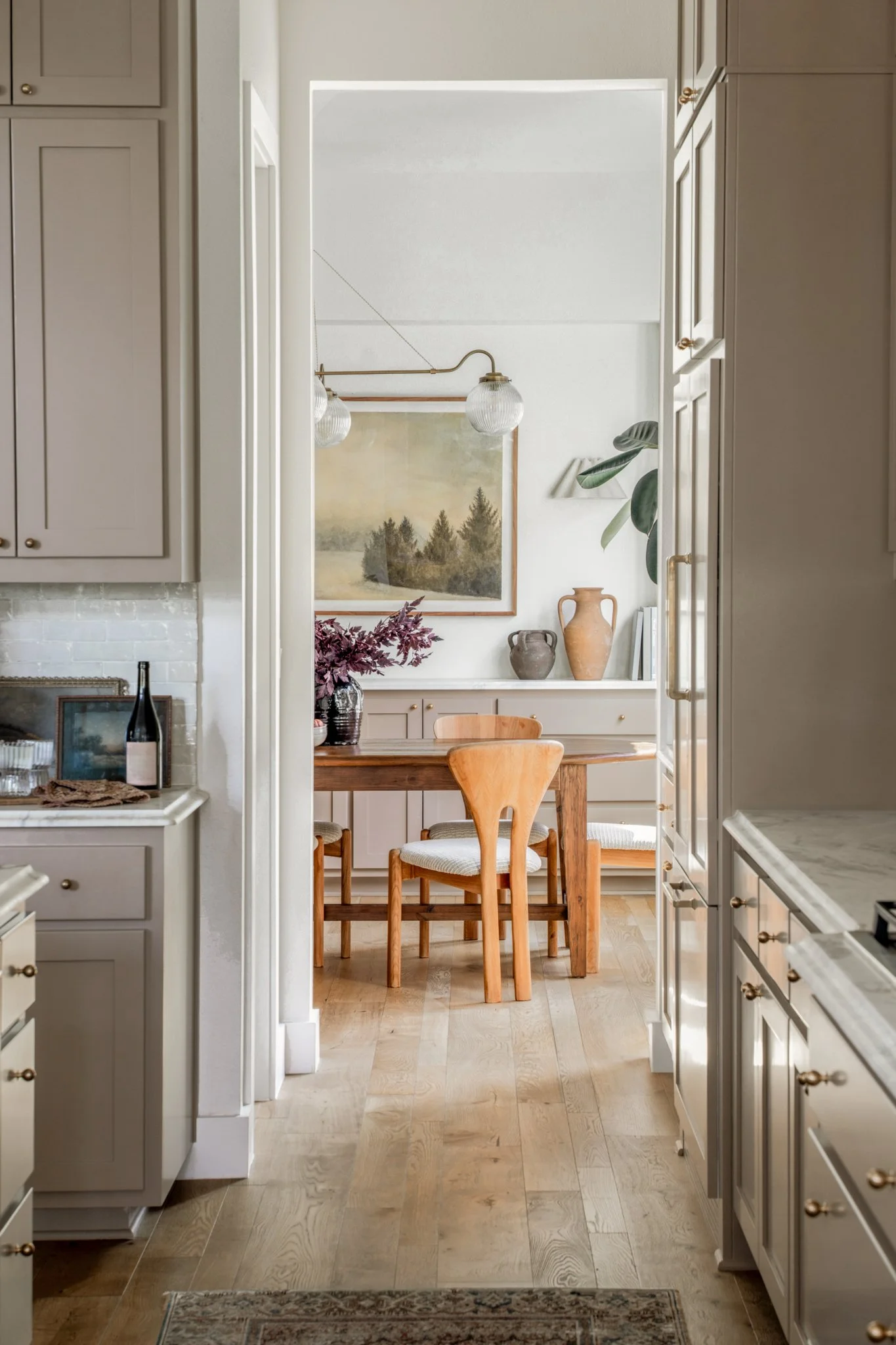 View through a doorway into a dining area with a wooden table and chairs, decorated with vases, artwork, and plants, in a modern, light-colored kitchen.