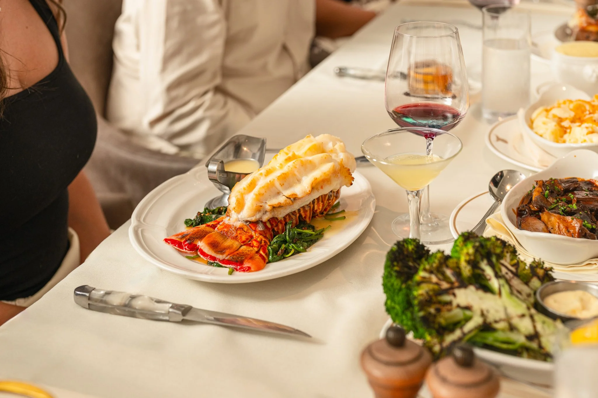 	
Restaurant Photography of Dean's Steakhouse at Kimpton Santo in San Antonio A plated lobster tail topped with mashed potatoes and gravy, served on a white plate with a side of greens, at a dinner table with wine, cocktails, and other dishes.