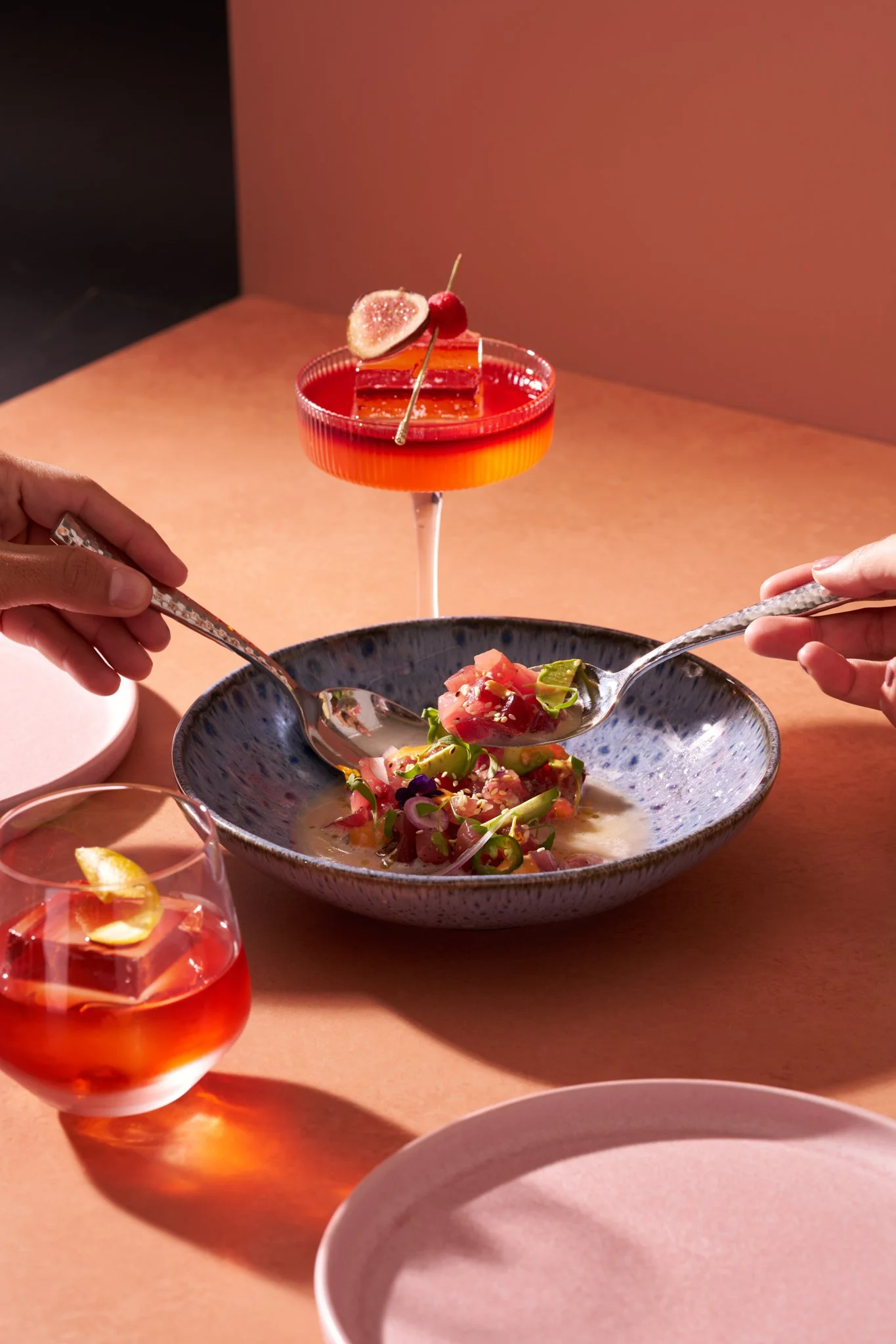 Restaurant photography  for Richard Sandoval Hospitality featuring People sharing a meal with a bowl of salad and two cocktails on a pink surface with a pink background.