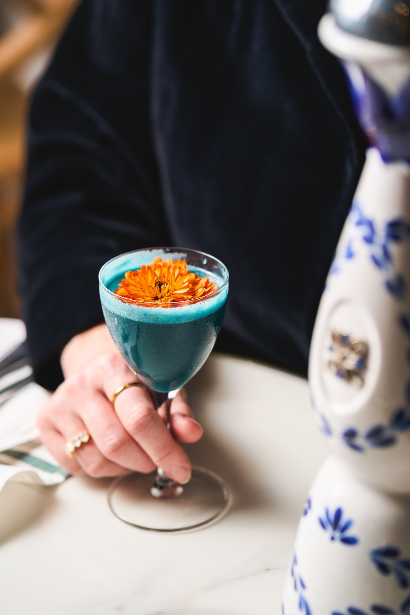 Beverage Photography at Blush Restaurant in San Antonio, Texas with A person holding a turquoise cocktail glass topped with an orange flower, sitting at a table with a white surface and a blue and white patterned cloth nearby.