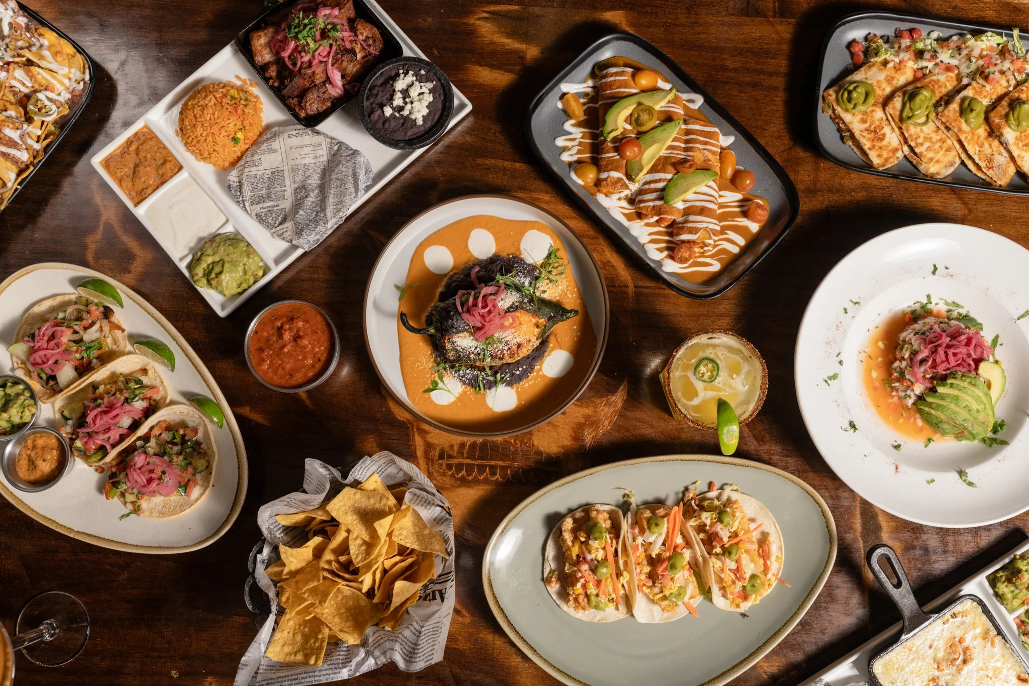 A variety of Mexican dishes on a wooden table, including tacos, grilled fish, chilaquiles, nachos, and a Mexican drink, with some chips and salsa.
