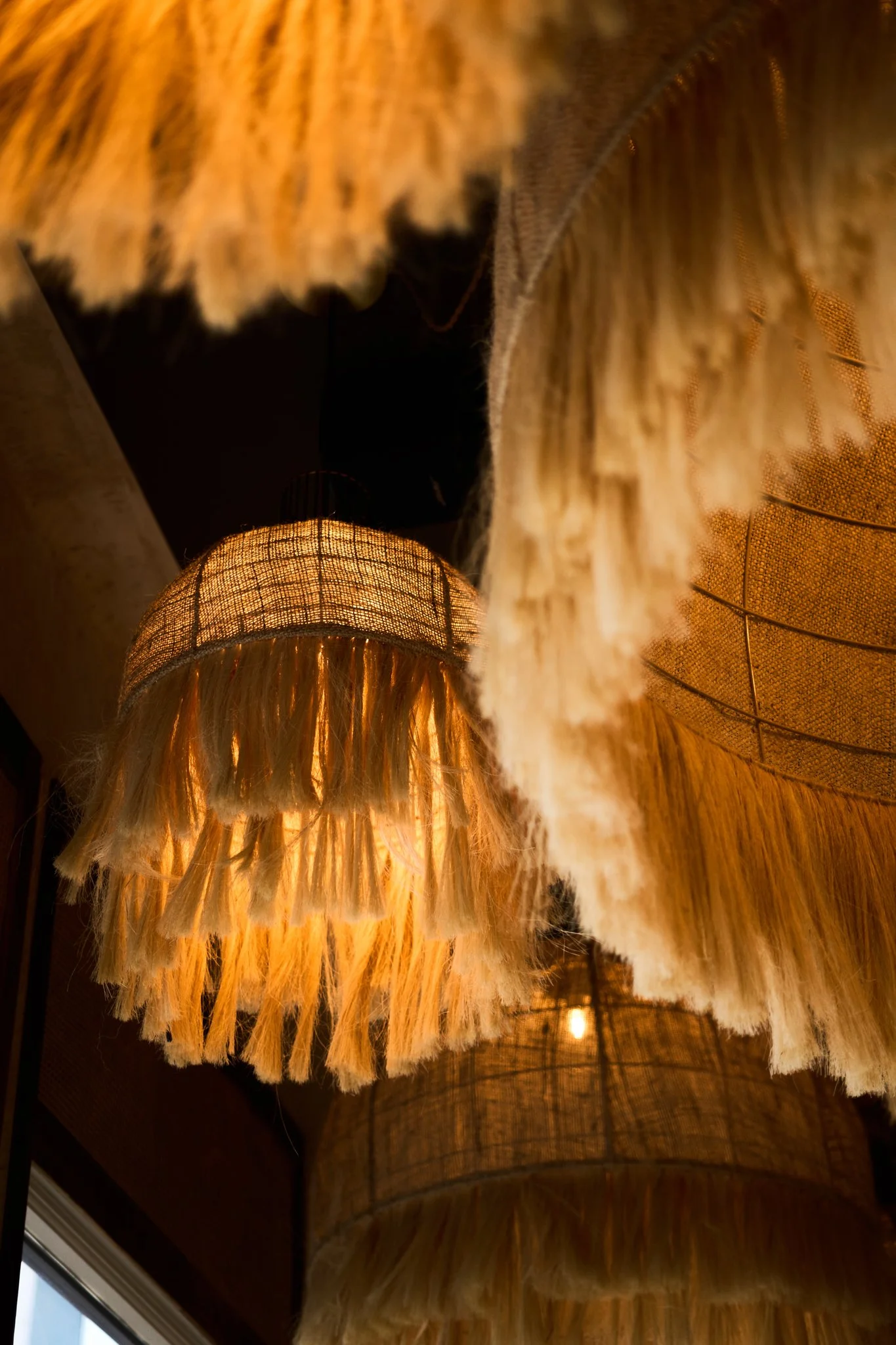 Hospitality Photography of Tamayo in Denver close-up of hanging decorative lampshades made from woven natural fibers, emitting warm light, with fringed edges.