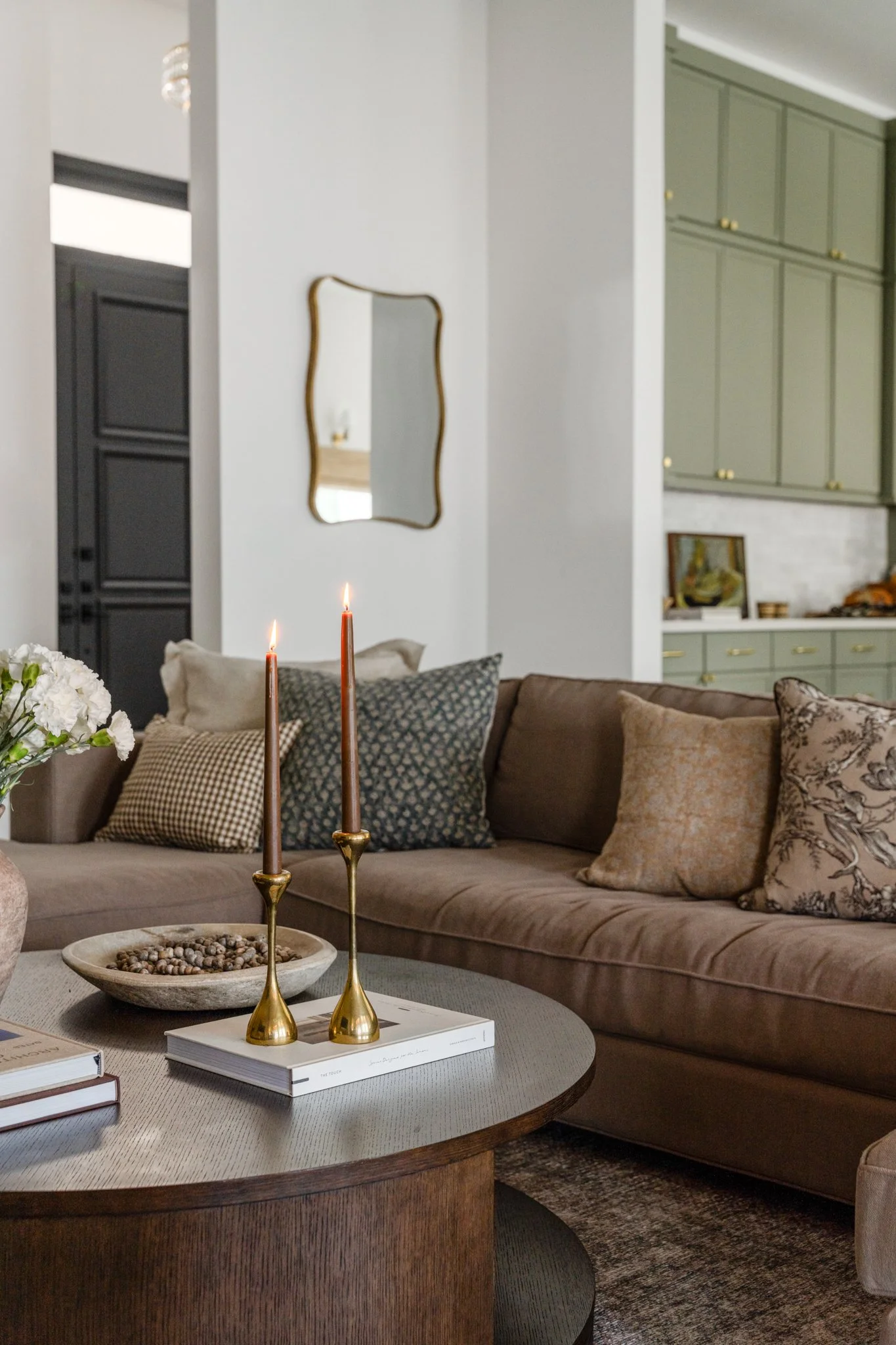 A cozy living room with a beige sectional sofa adorned with patterned and solid cushions. A round wooden coffee table in front holds two lit brown candles in gold candle holders, a bowl of small stones, and a book. In the background, a mirror with a 