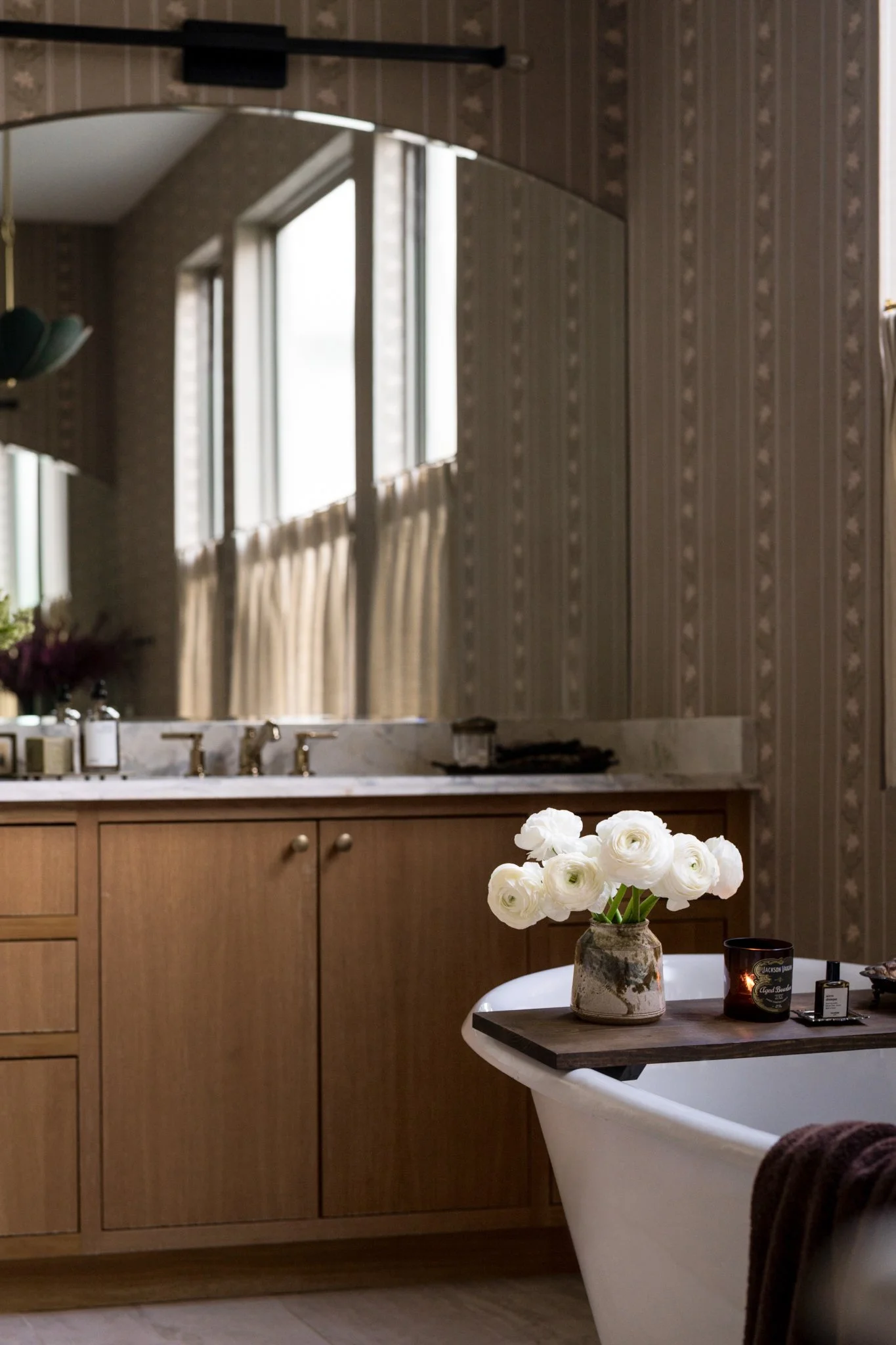 A bathroom with a wooden vanity, a marble countertop, a large mirror, and windows with beige curtains. There is a bathtub with a wooden tray holding white flowers, a candle, and toiletries.