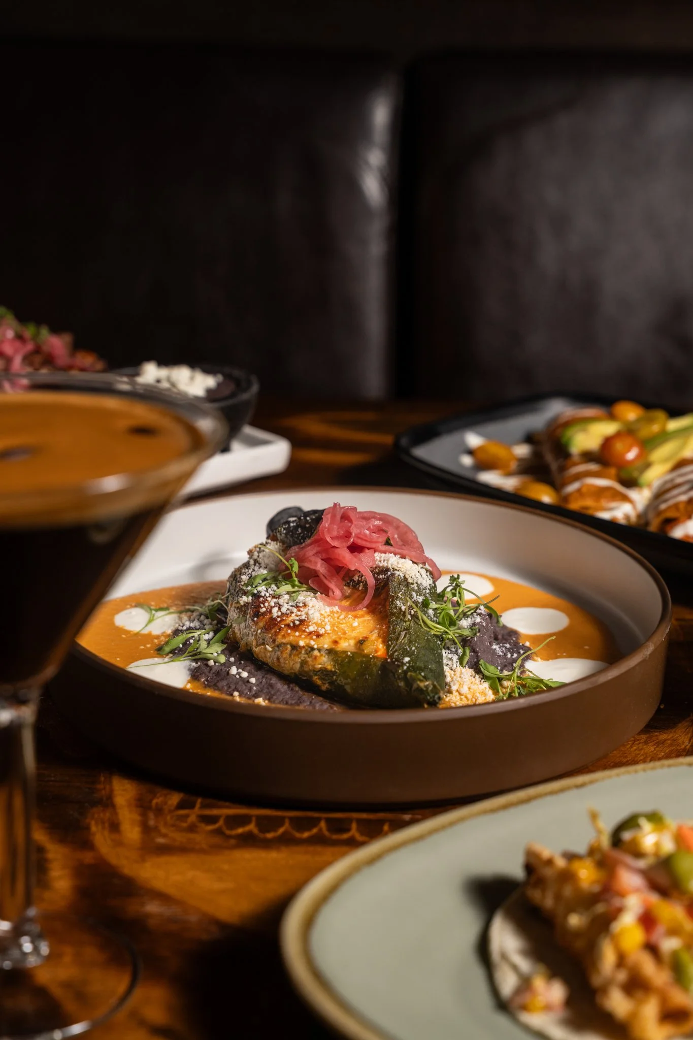 Food photography for Richard Sandoval of serving of stuffed poblano topped with pink pickled onions, garnished with herbs, on a dark round plate with a drizzle of white sauce, surrounded by other dishes and a glass of dark beverage on a wooden table.