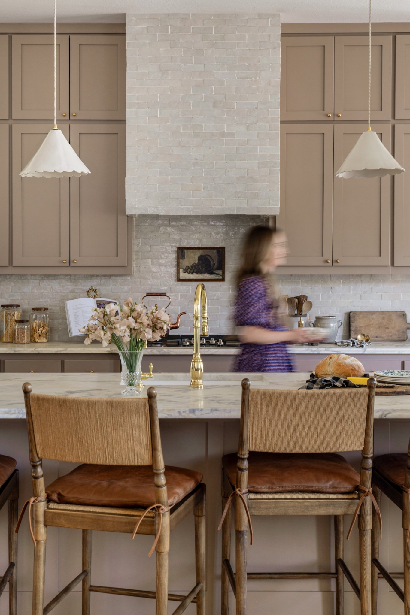 A woman in a purple dress in a modern kitchen with beige cabinets, a white brick backsplash, a marble island counter, gold faucet, a vase of flowers, and bread on the counter. Two white pendant lights hang above.