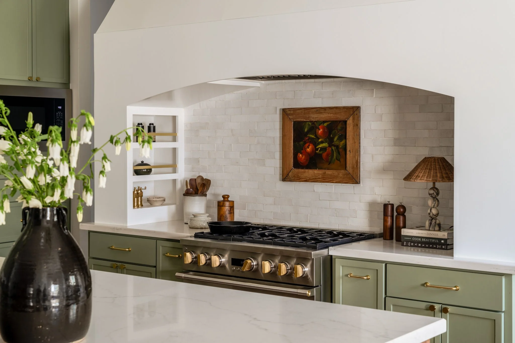 A modern kitchen with a white brick backsplash, a stainless steel stove, and green cabinets with gold handles. Decor includes a framed painting of peaches, a table lamp, and books.